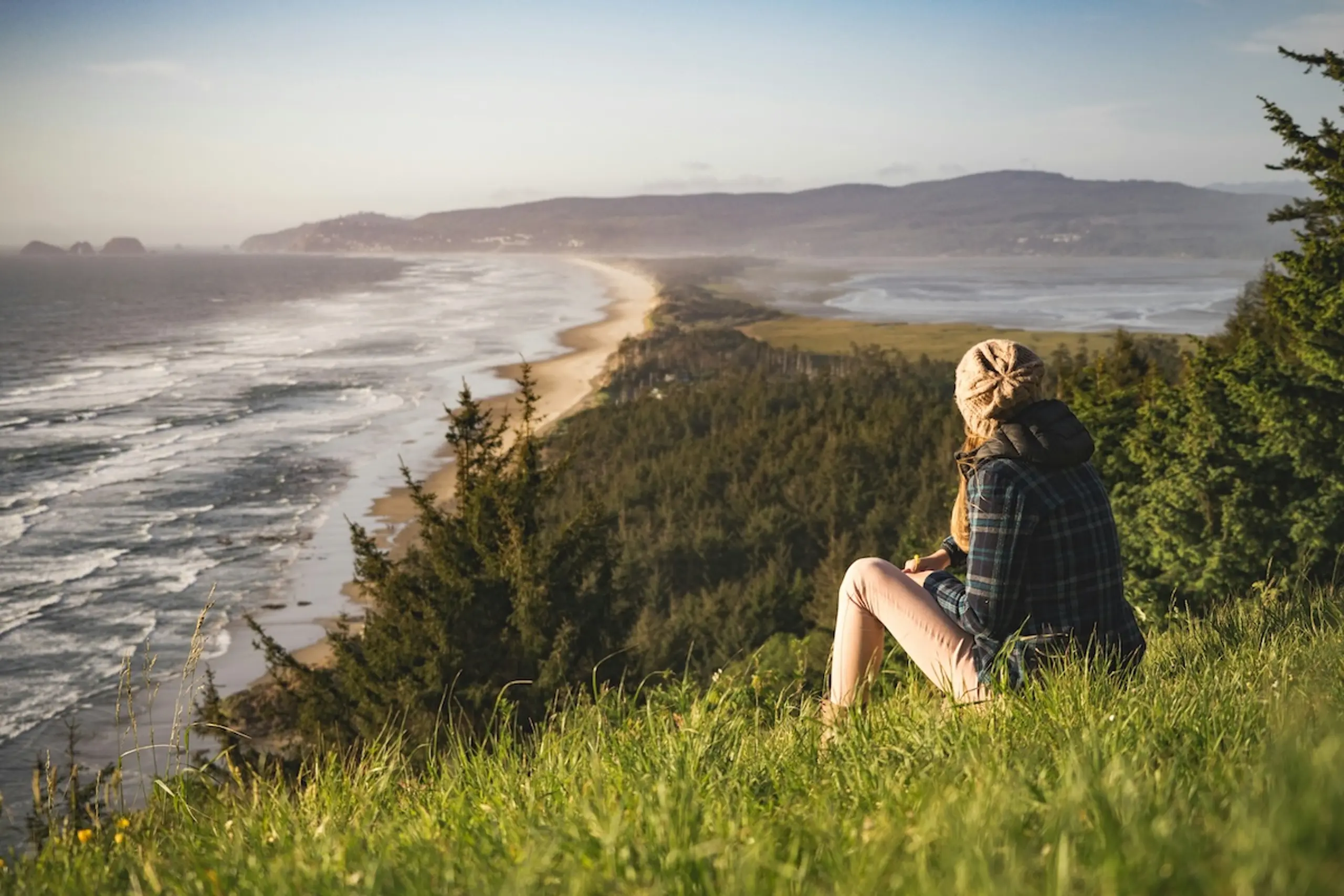 person wearing hat looking onto beach and mountains from a cliffside