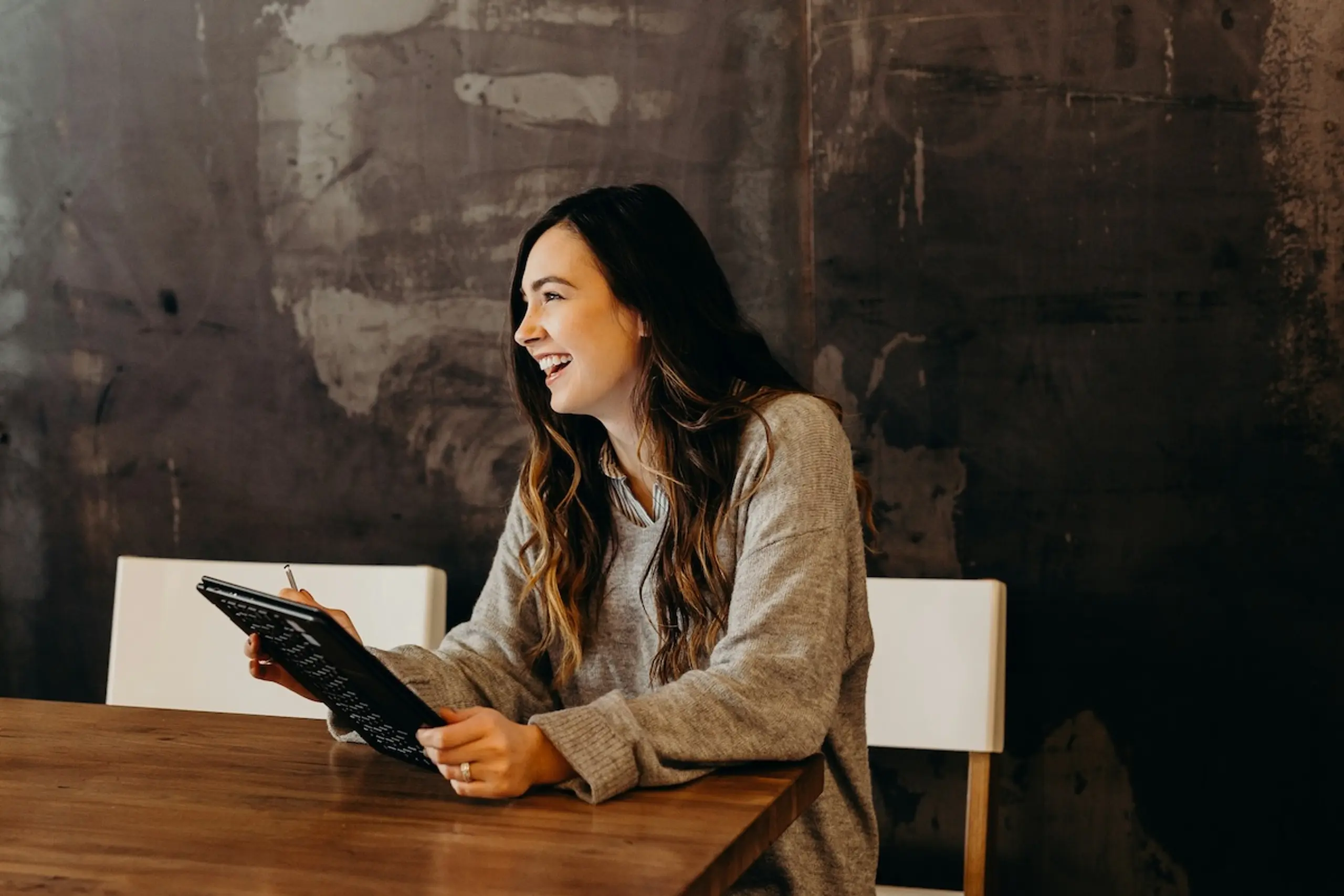 woman smiling while holding notebook seated at a table in front of a grey wall
