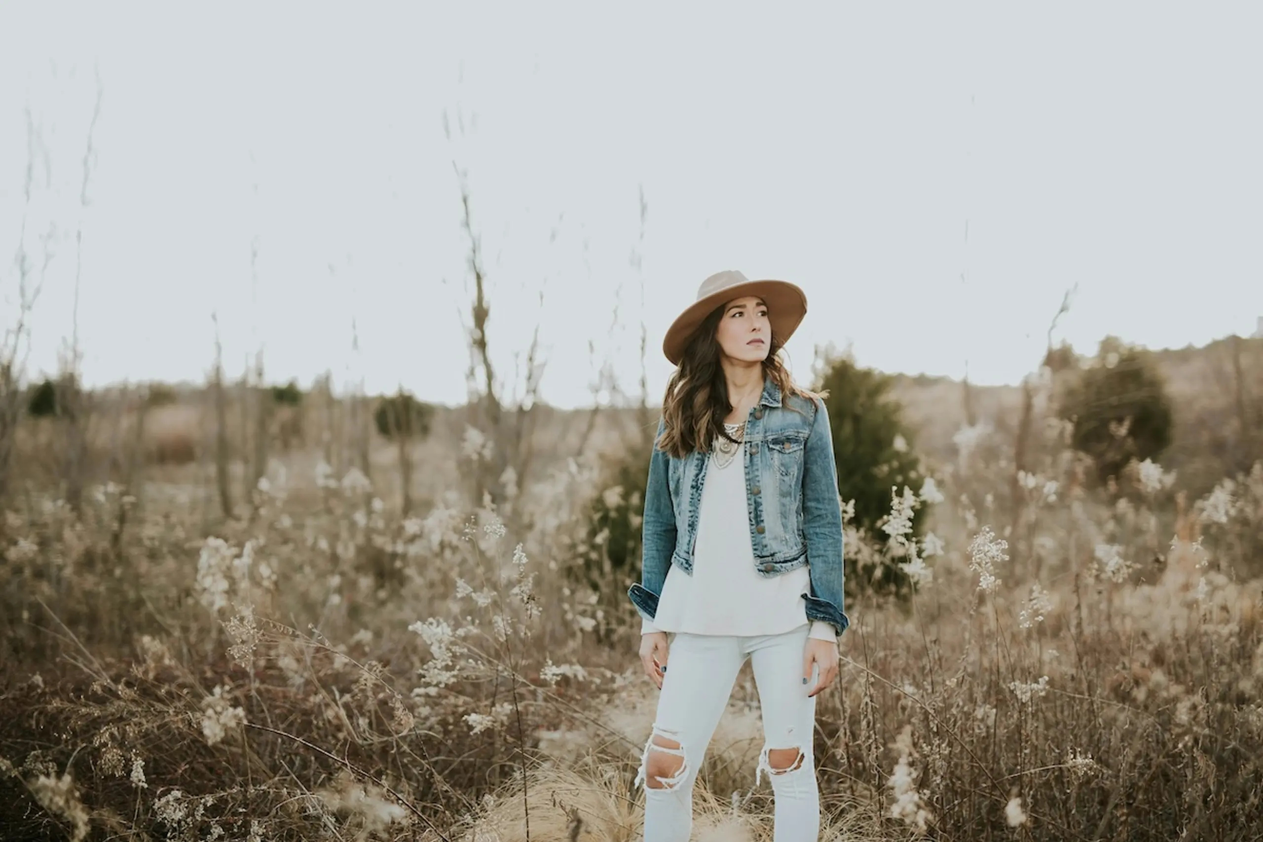 woman standing in field wearing hat, jean jacket, and white shirt and jeans
