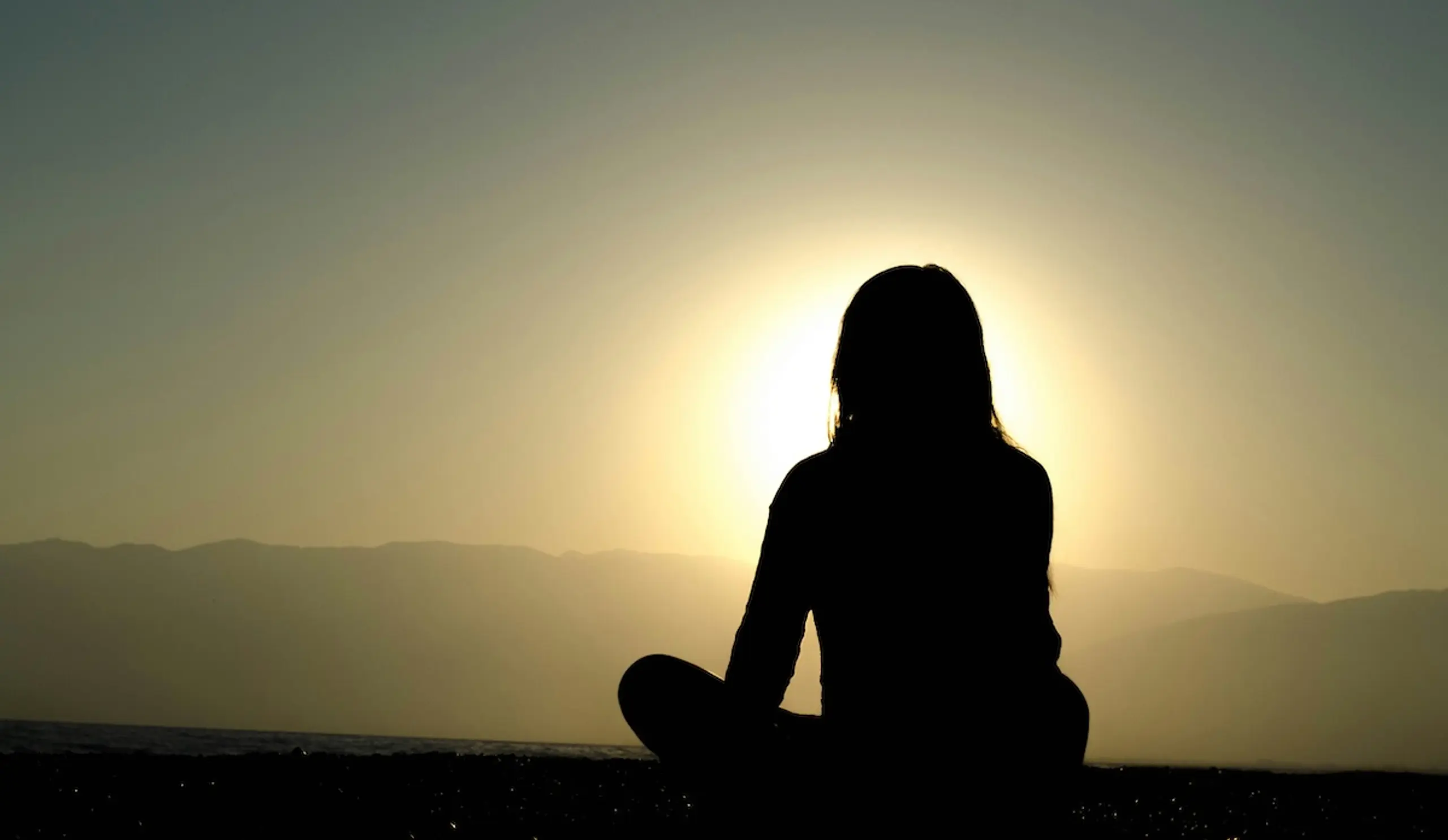 person sitting with crossed legs watching sunrise over mountains in the distance
