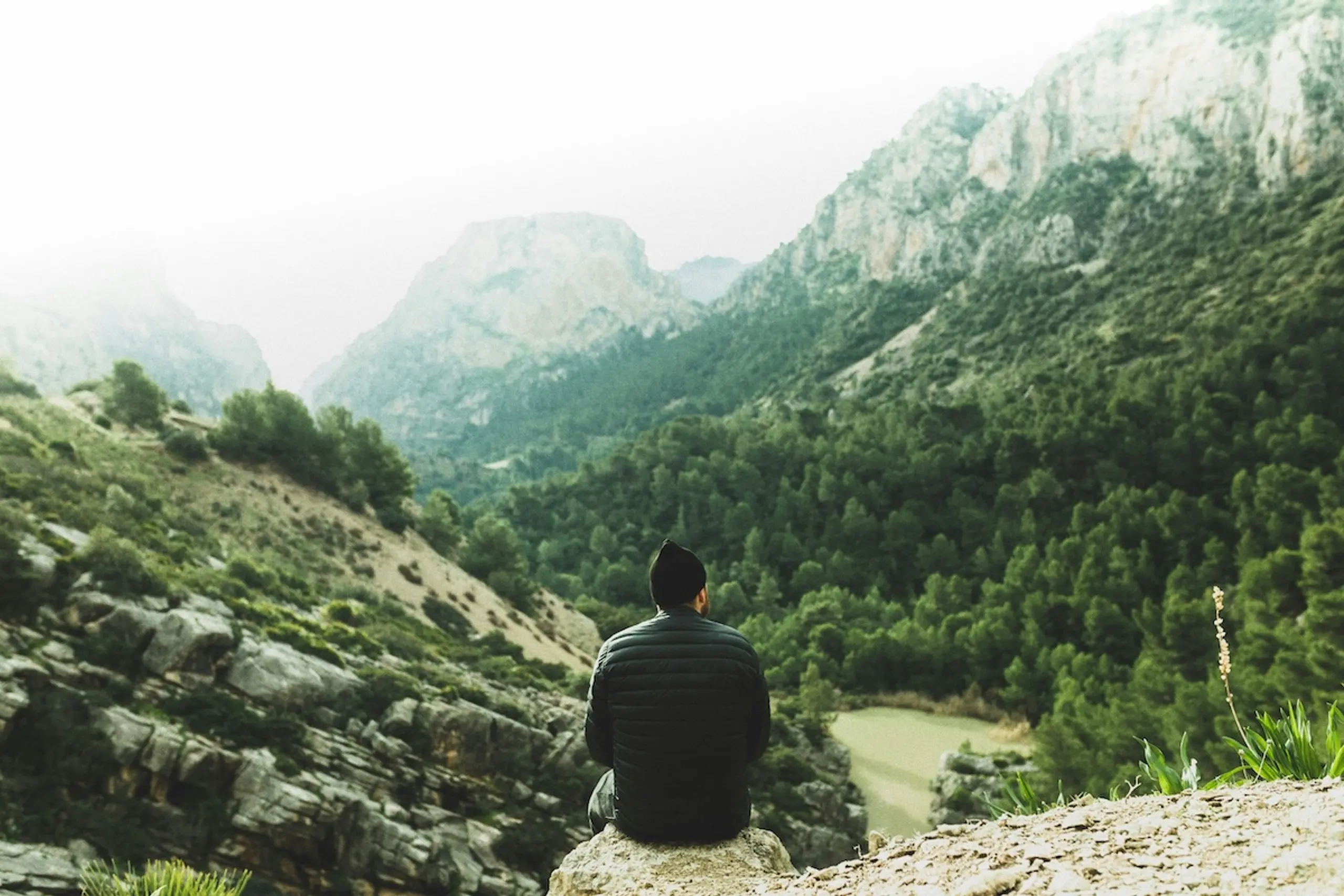 person in hat and jacket sitting on a rock looking over mountains and trees in a valley
