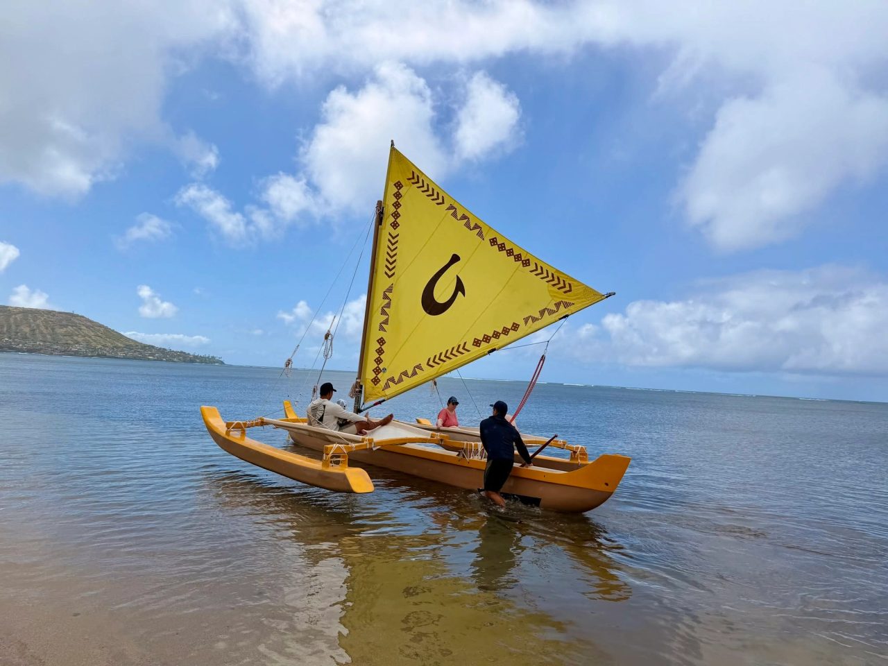 People preparing a traditional yellow-sailed canoe on the shore in Hawaiʻi