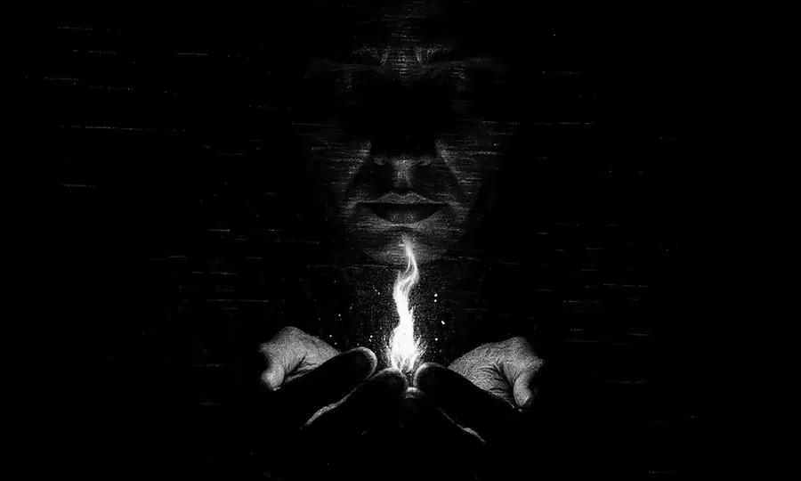 a man cups a little light in complete darkness with his face barely seen in the shadows