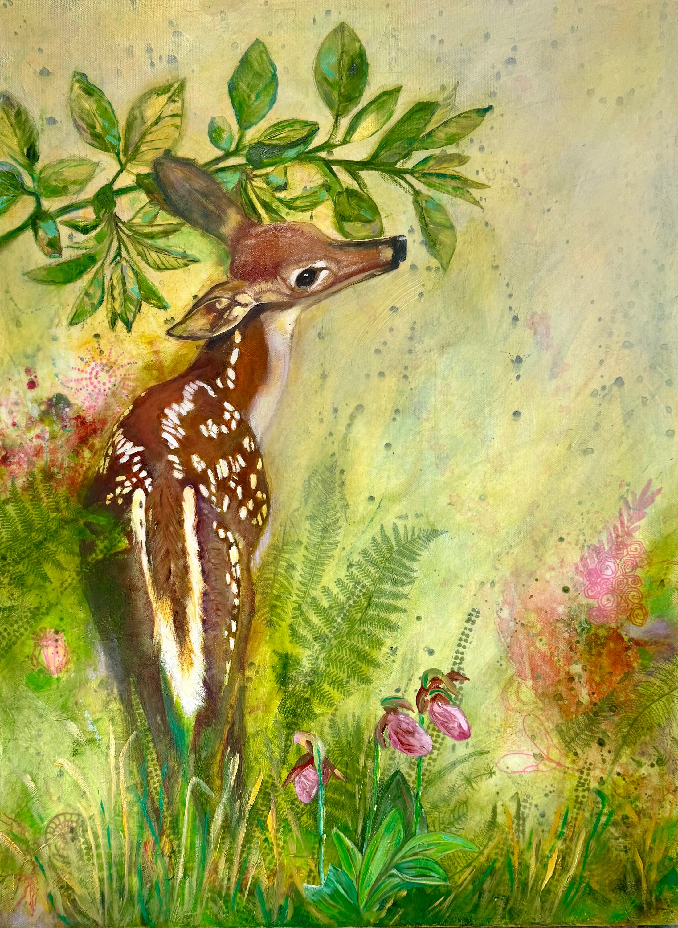 Spotted fawn standing beneath leafy branches amid ferns and pink woodland flowers.