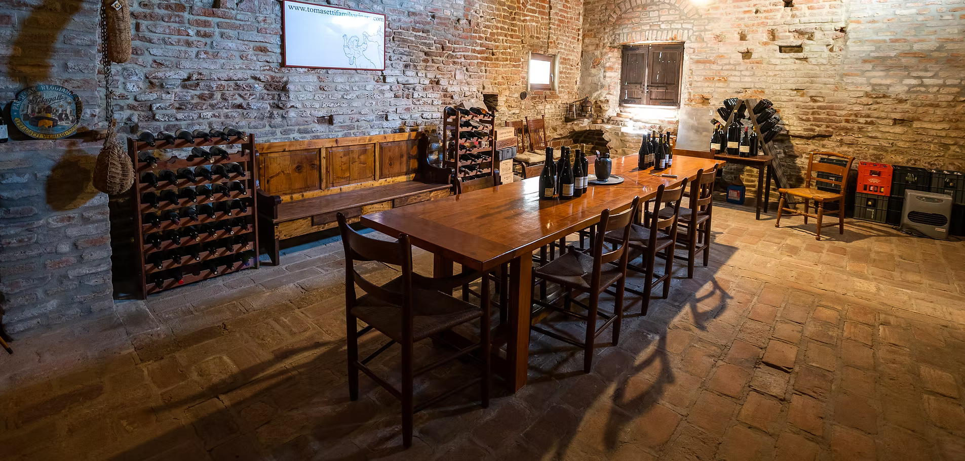 A rustic wine cellar with exposed brick walls, a long wooden table set with wine bottles, and wine racks.
