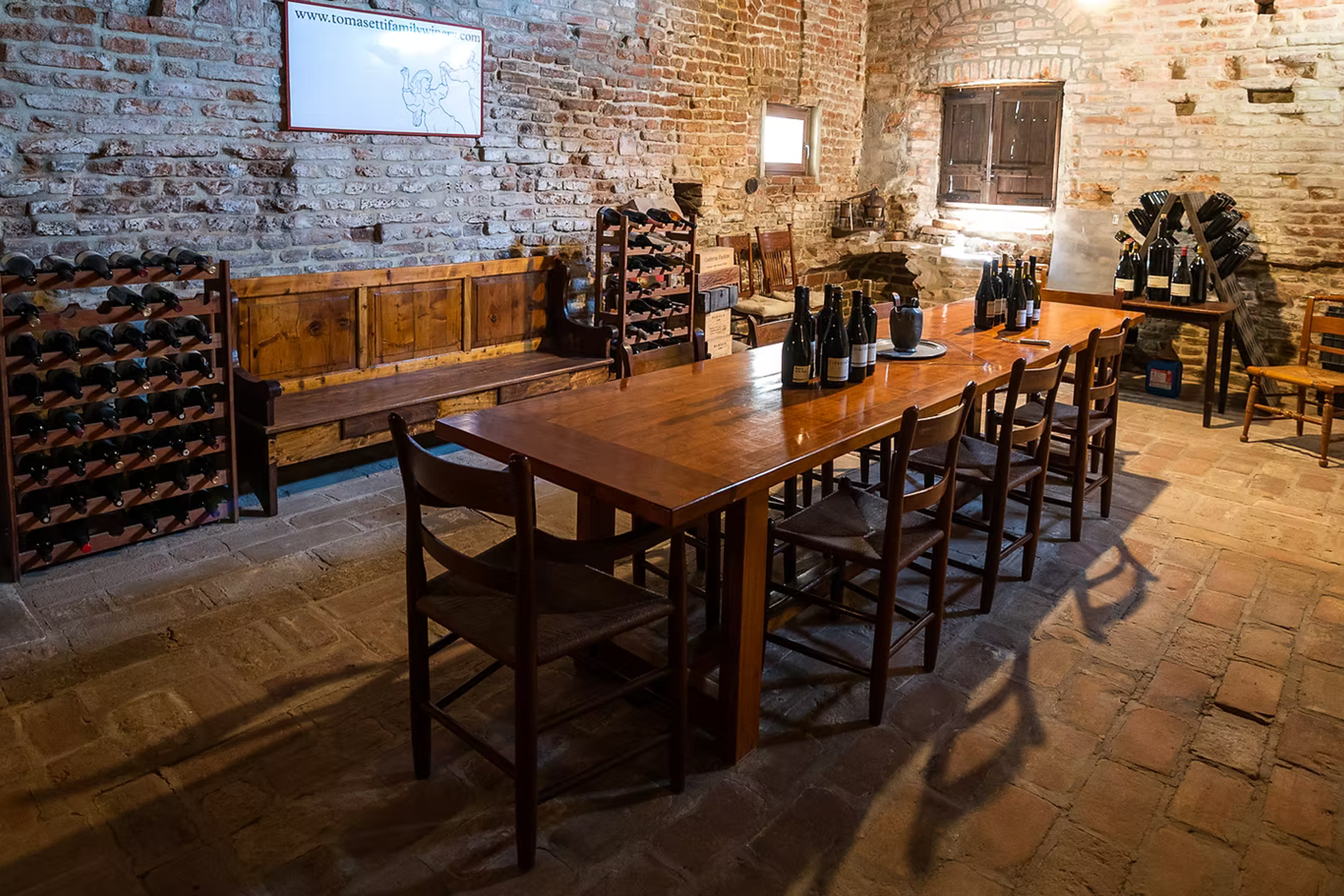 A rustic wine cellar with exposed brick walls, a long wooden table set with wine bottles, and wine racks.