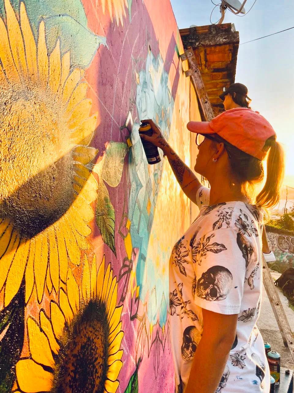 Tuane Arte pintando mural de girassol com spray ao pôr do sol