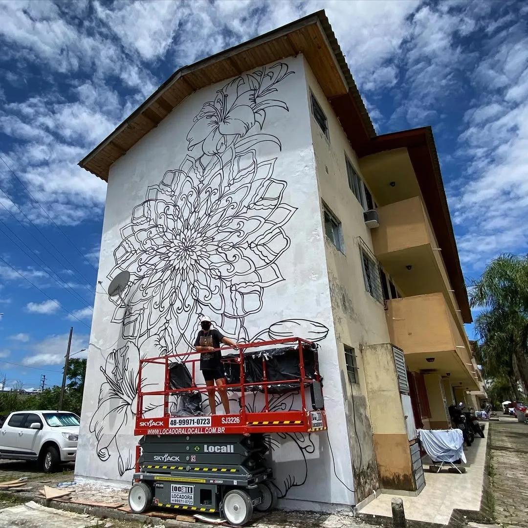 Tuane Arte pintando o contorno de um mural floral em prédio