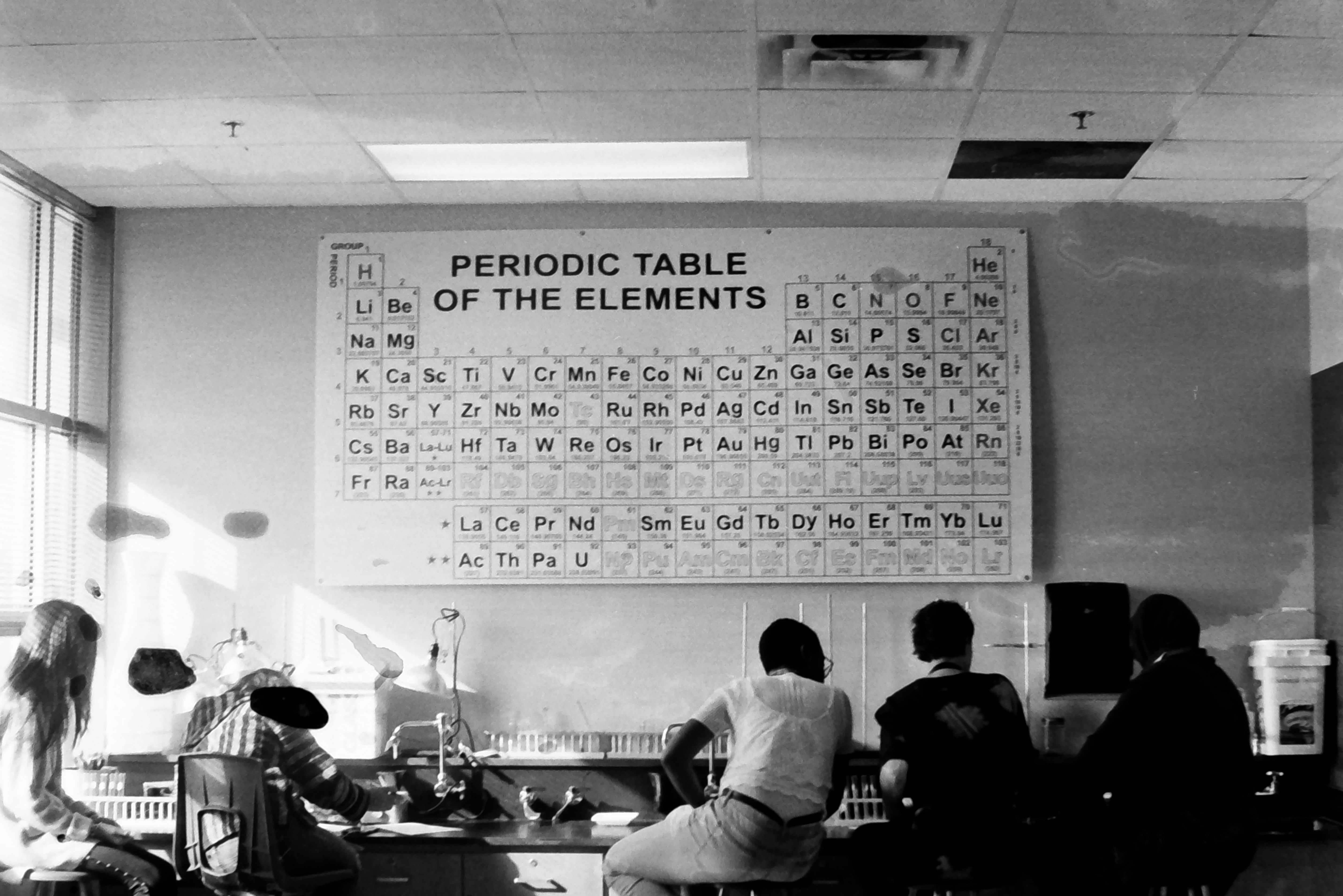 A chemistry lab with a large Periodic Table on the wall, where students are seated and conducting experiments. A chemistry lab with a large Periodic Table on the wall, where students are seated and conducting experiments.