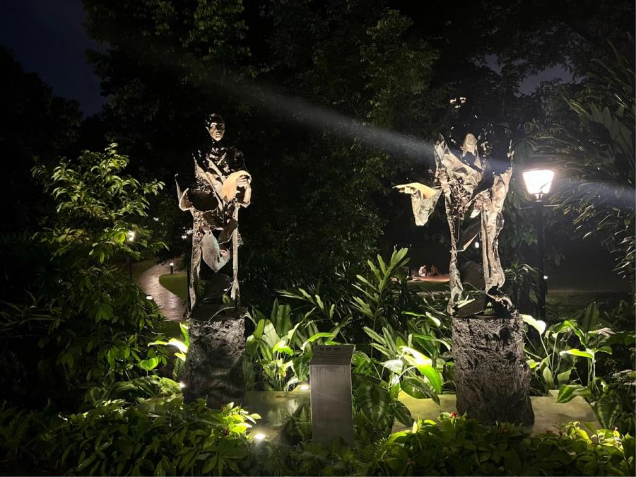 Two bronze sculptural figures of Stamford Raffles and Nathaniel Wallich lit by ground spotlights in a lush nighttime garden at Fort Canning Park