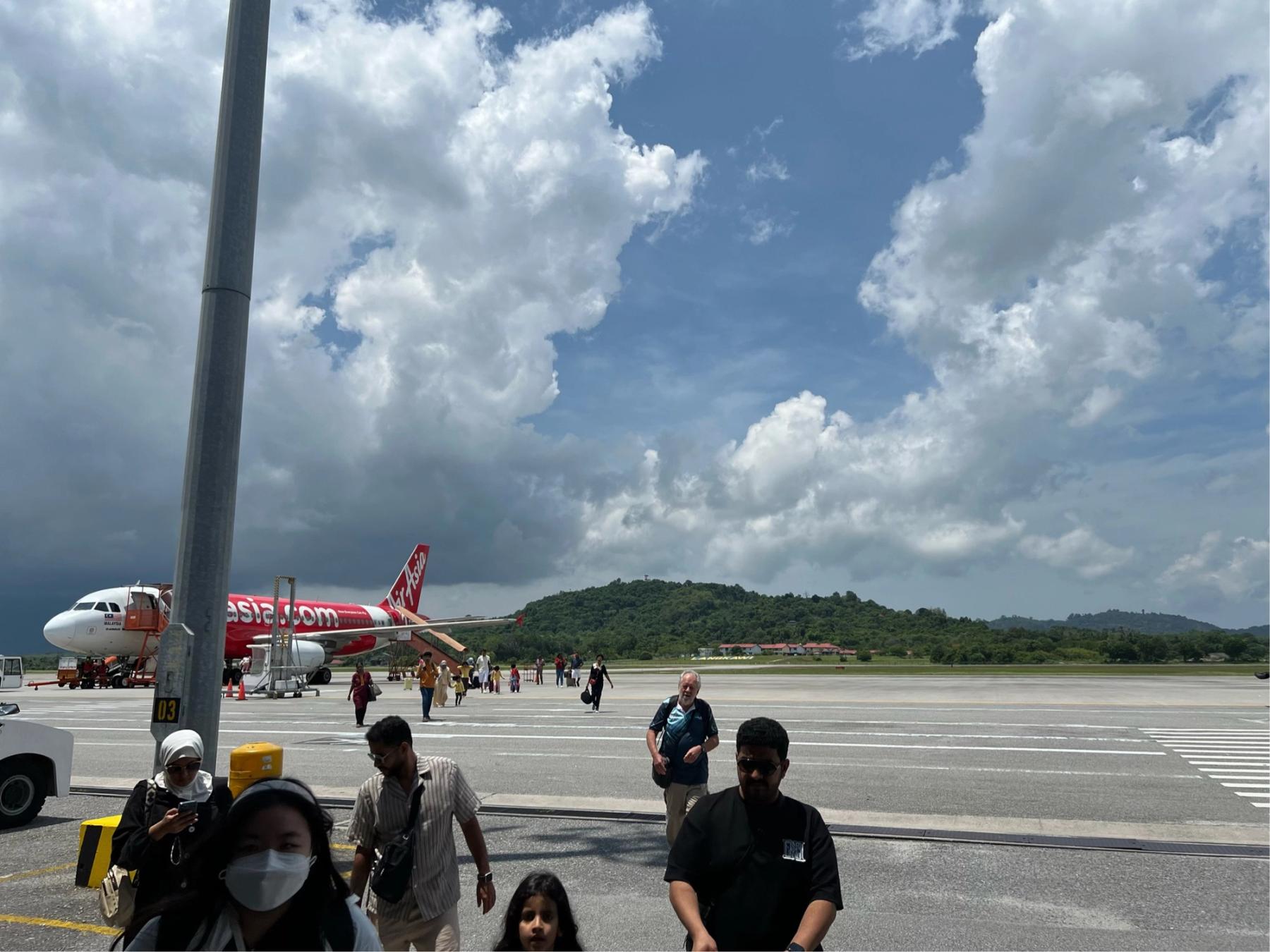 Passengers walking across the open tarmac at Langkawi Airport, an AirAsia jet parked behind them beneath dramatic tropical clouds and a forested hill