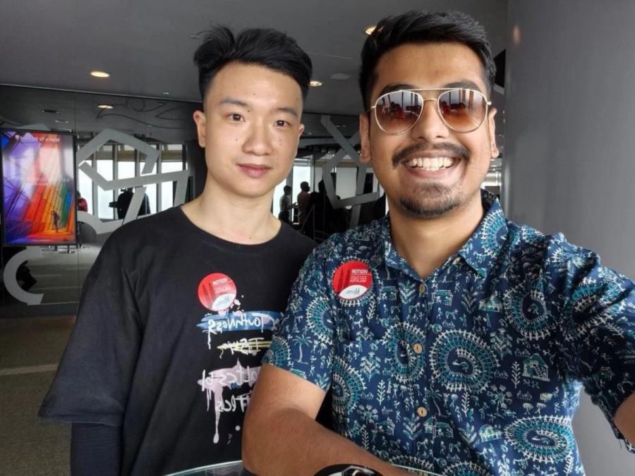 Two men smiling side by side inside the Petronas Twin Towers observation area, both wearing red entry badges