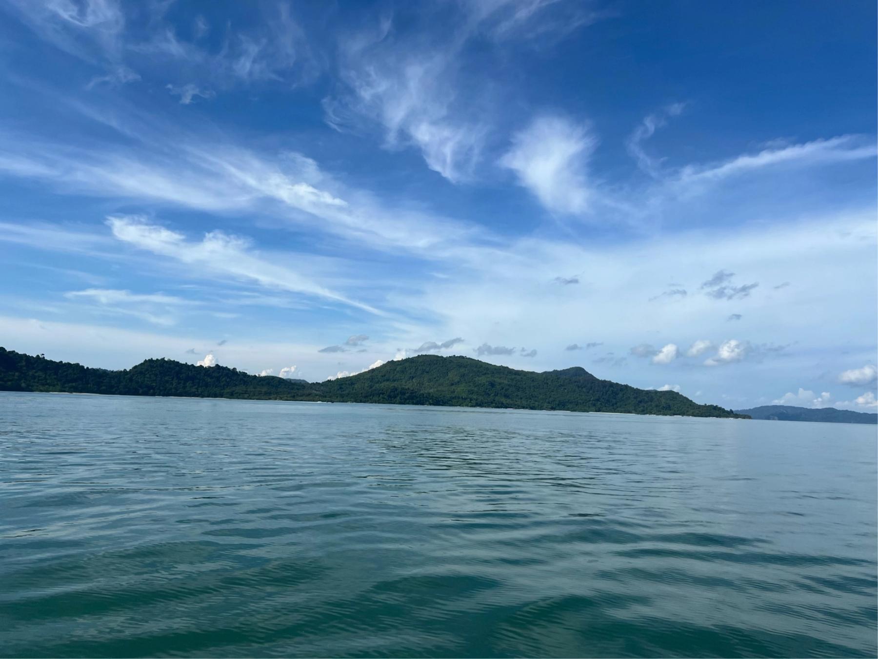 Turquoise sea stretching toward a densely forested island under a wide blue sky with wispy clouds, viewed from a boat