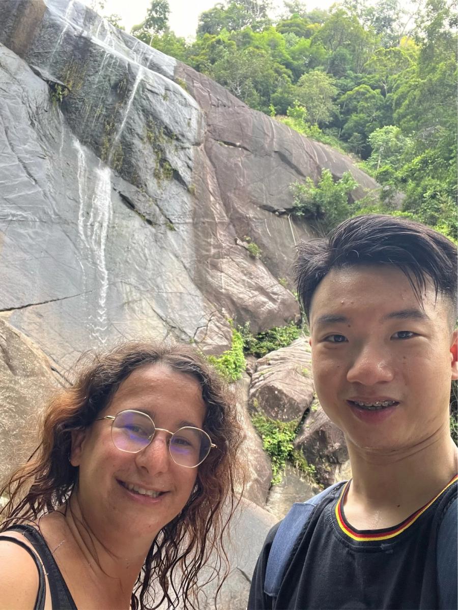 Two travelers smiling in a selfie in front of a large granite rock face with thin waterfalls trickling down, dense jungle behind them