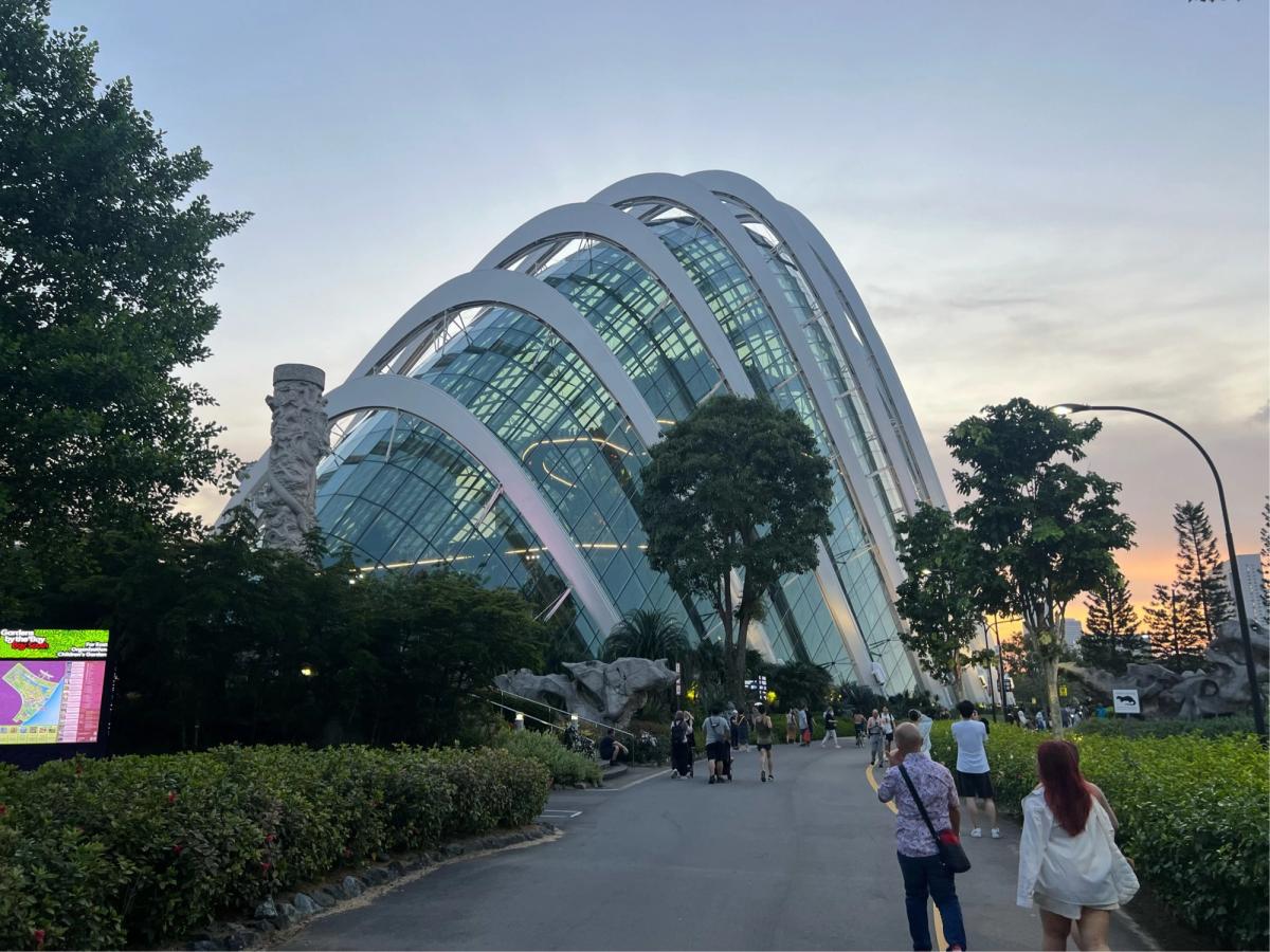 Flower Dome conservatory at Gardens by the Bay at dusk, its white arched ribs glowing against a pink and blue sky, visitors walking the path toward the entrance