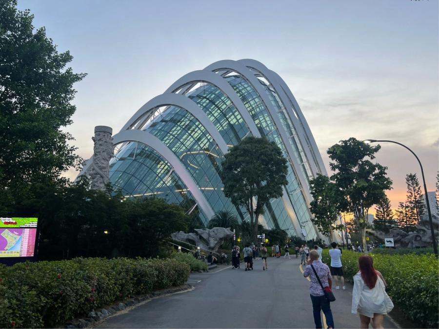 Flower Dome conservatory at Gardens by the Bay at dusk, its white arched ribs glowing against a pink and blue sky, visitors walking the path toward the entrance