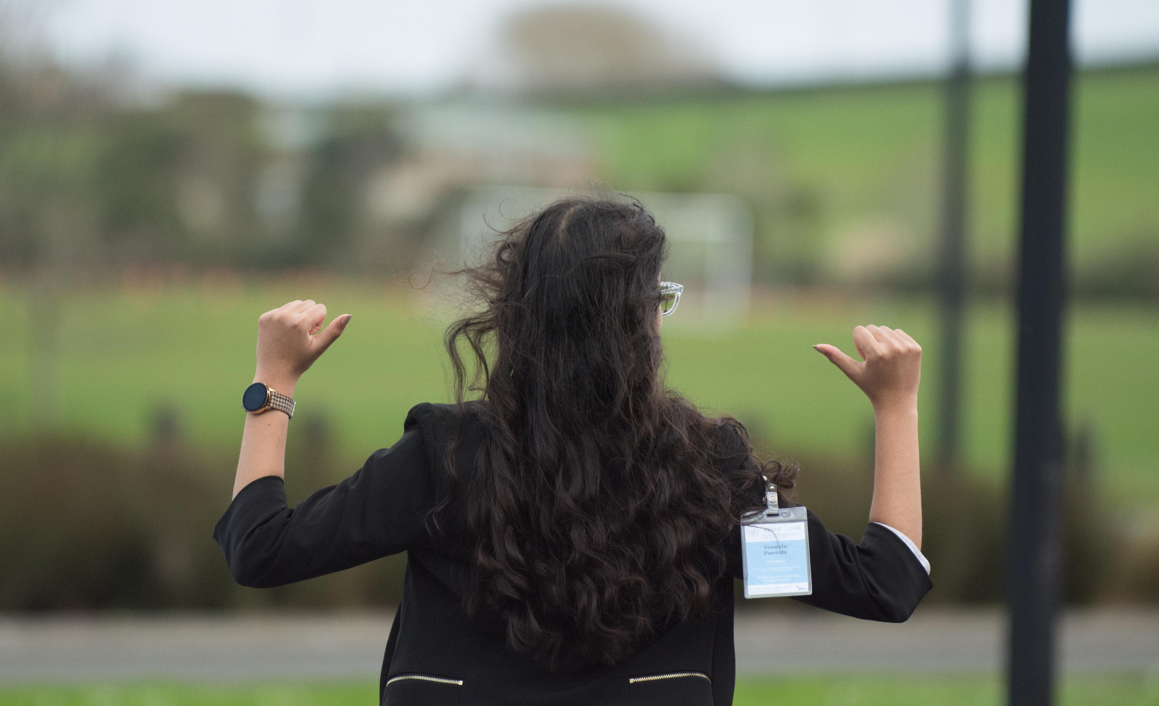 A person with long dark hair, seen from behind, points their thumbs towards themselves outdoors.