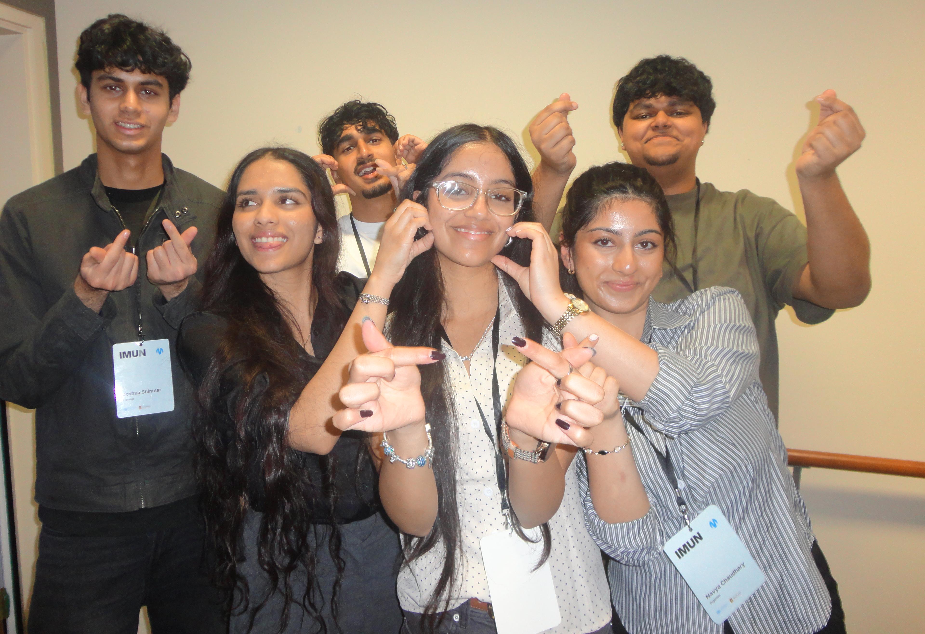 A person with long dark hair, seen from behind, points their thumbs towards themselves outdoors.