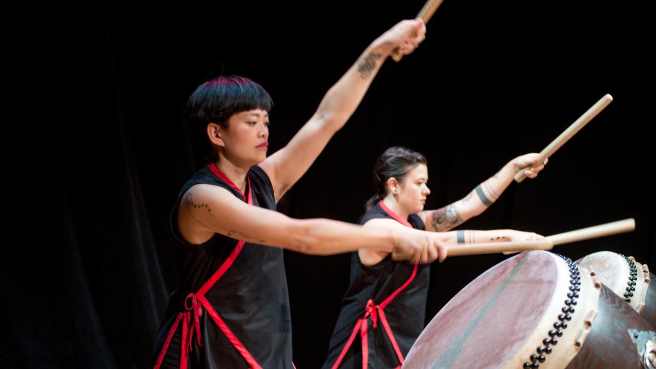 Two women hold drumsticks suspended above their taiko drums in a mid-performance pose.