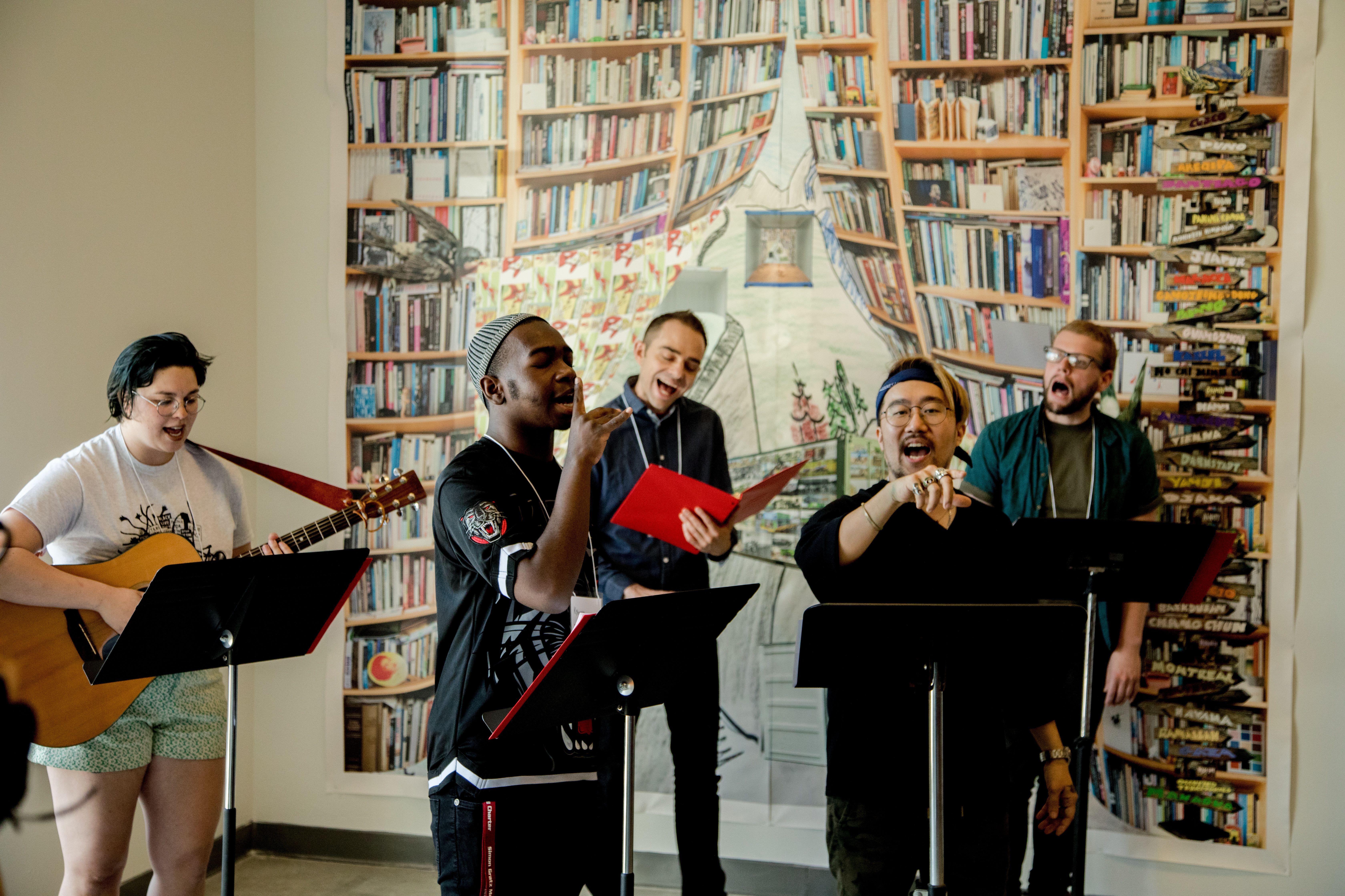 A band of five singing and performing in front of a painting of bookshelves.