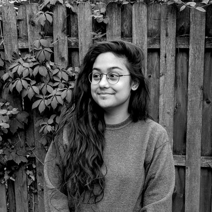 Black and white headshot of Marria Nakhoda in front of a wood fence with leaves sticking out of it.