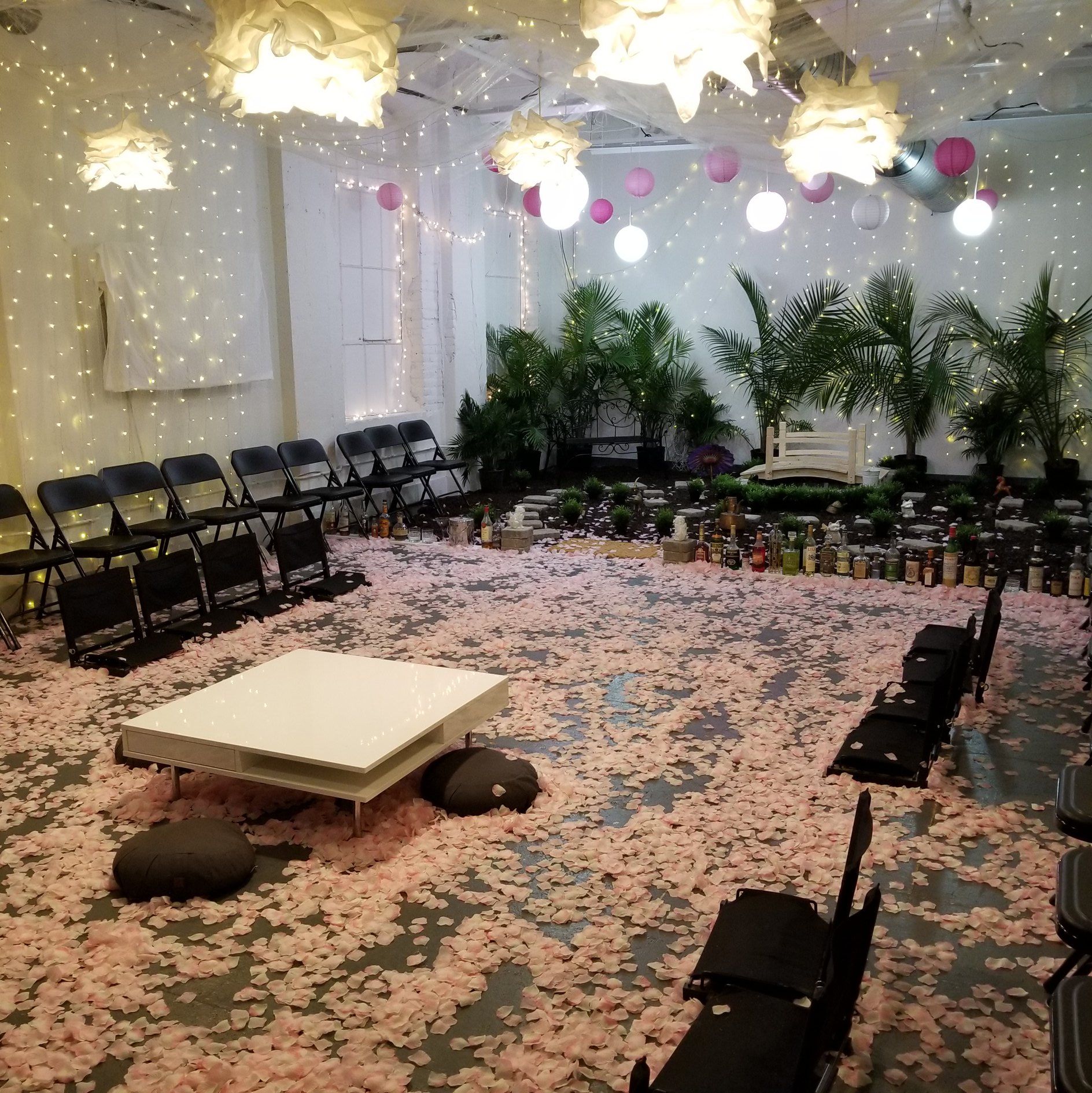 Room with chairs decorated with lights, balloons, small trees, candles, and flower petals all over the floor.