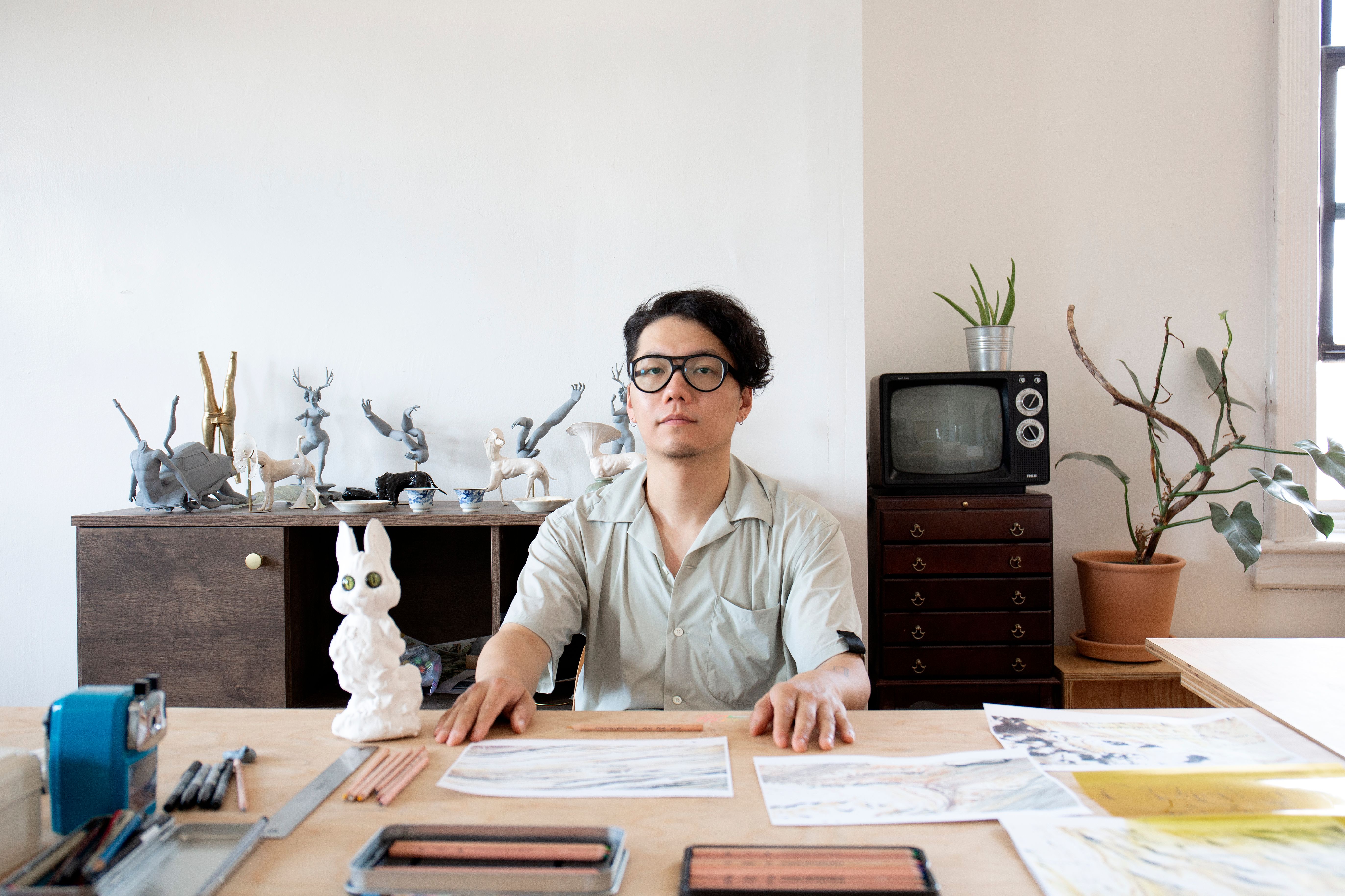 Artist Frank Wang looking into the camera, sitting infront of a desk, many papers and stationary on the desk, many figurines on a shelf in the background on the left, TV and plant in a pot on the right