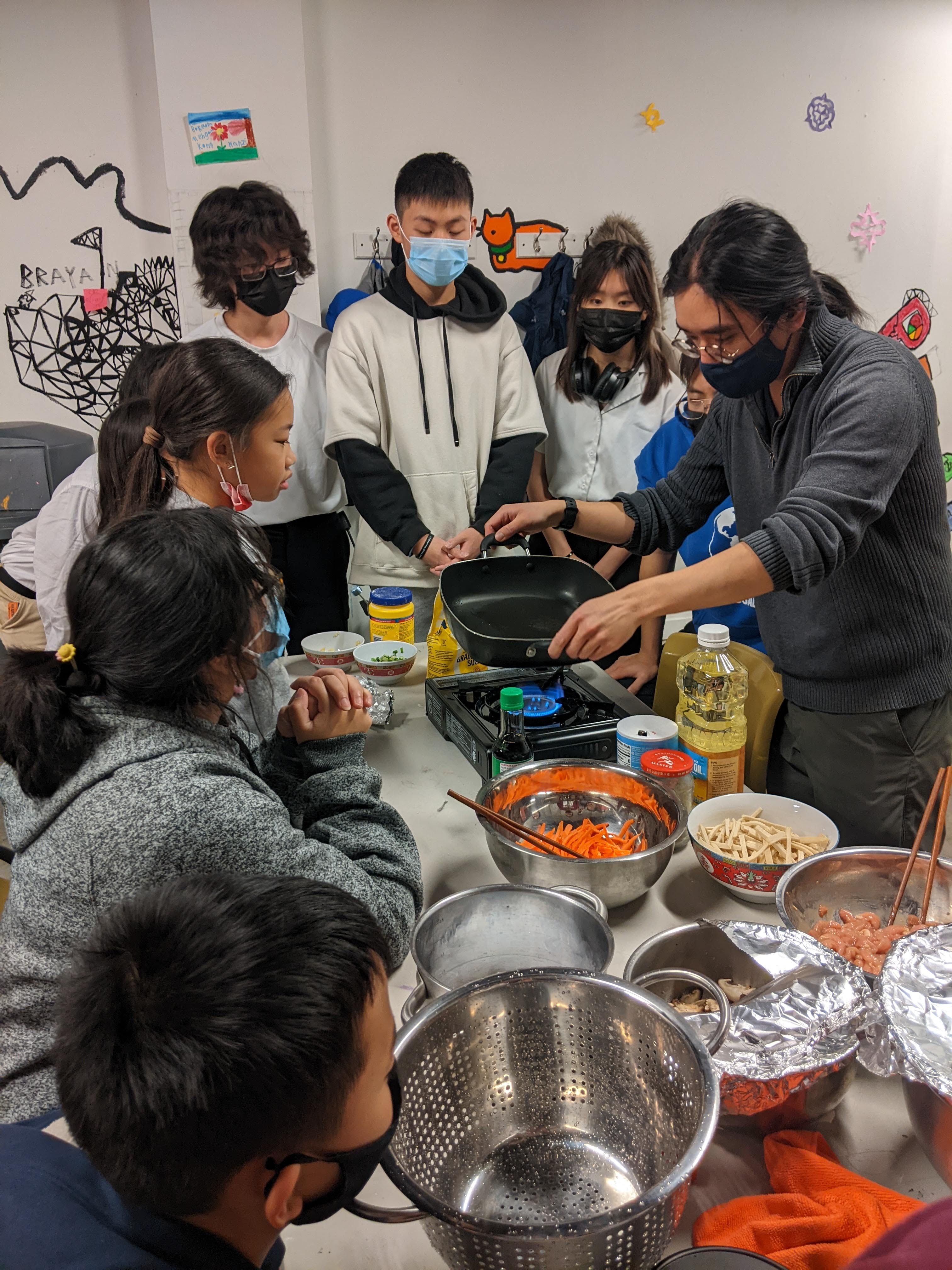 Students from Youth Art Workshop gathered around and watching a chef cook.