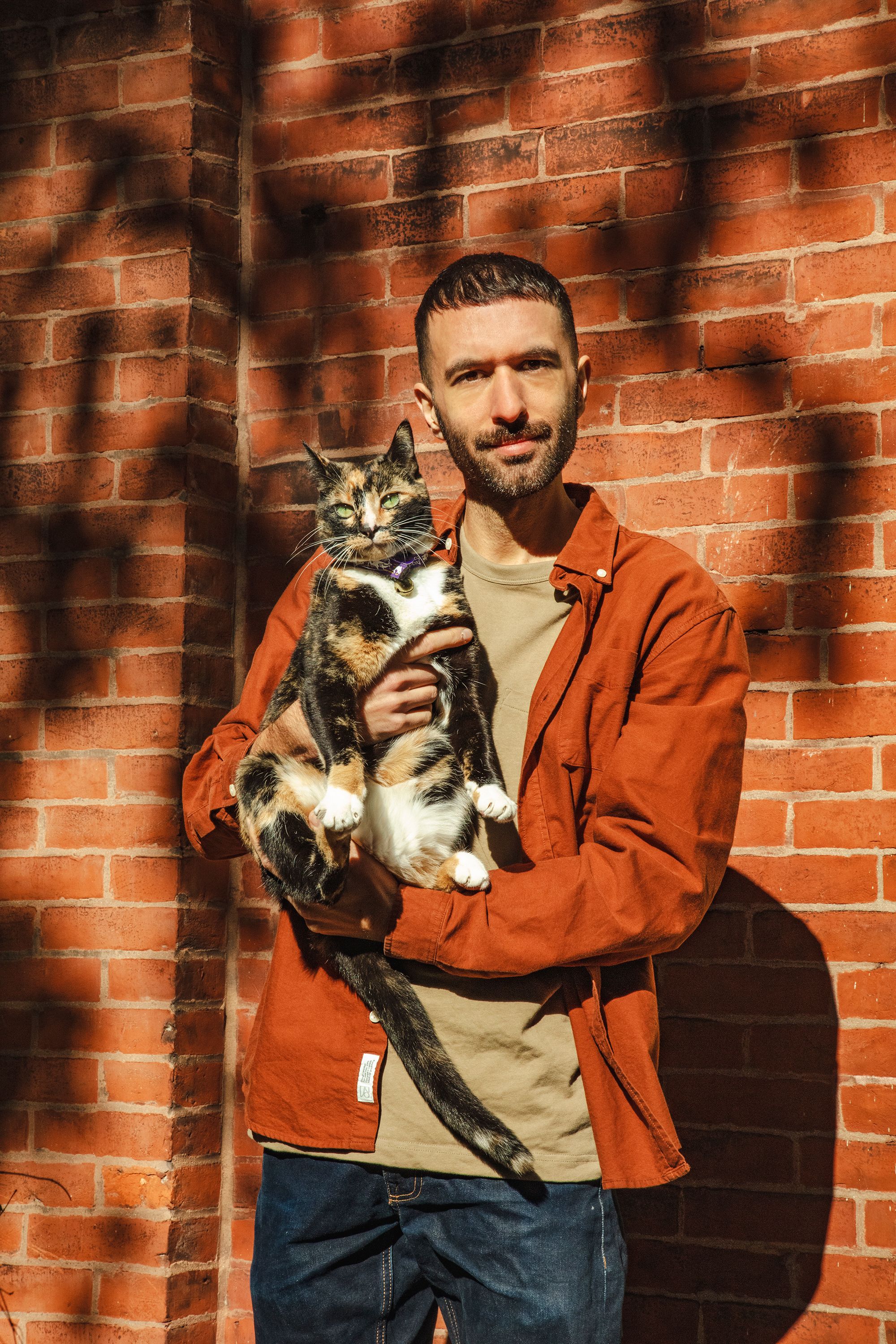Artist Rami George holding a cat, posing for a headshot photo, standing in front of red bricked wall