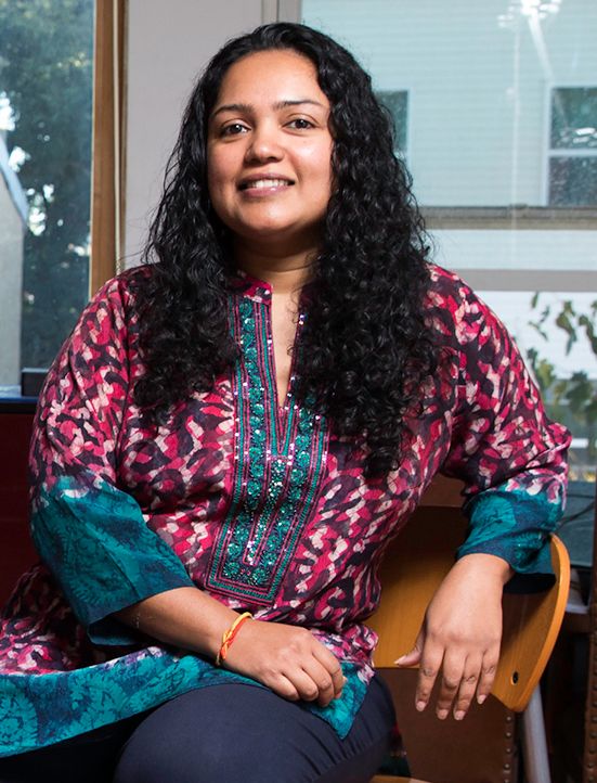 Artist Anula Shetty sitting for a portrait, with a colorful shirt and smiling, in front of a window