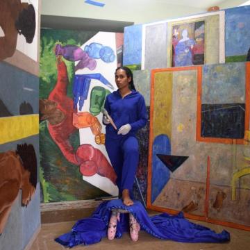Mequitta Ahuja holding a paintbrush while stepping on a stool in front of paintings.