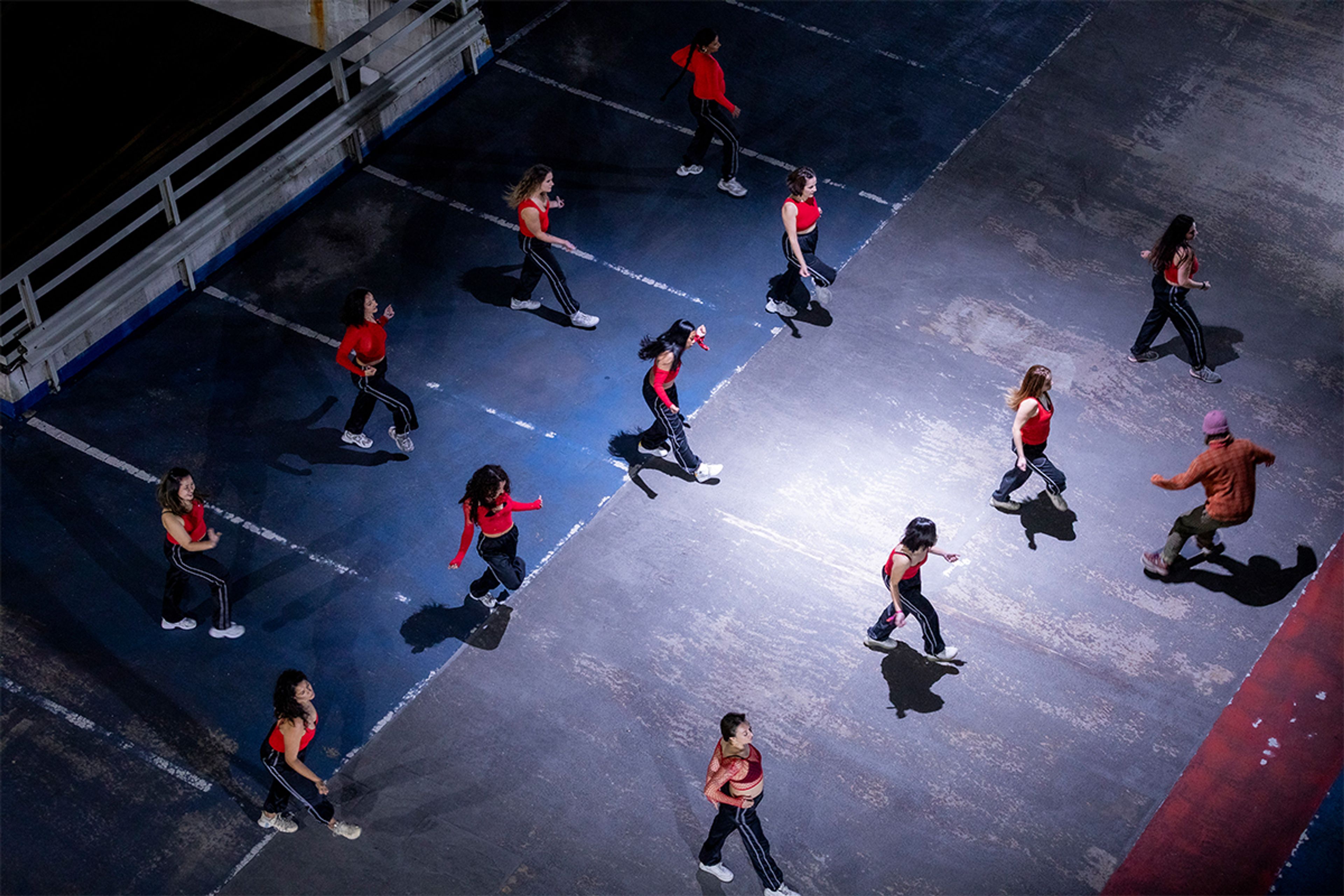 A group of people dressed in matching tracksuits dance in formation within a car park lit by street light at night.