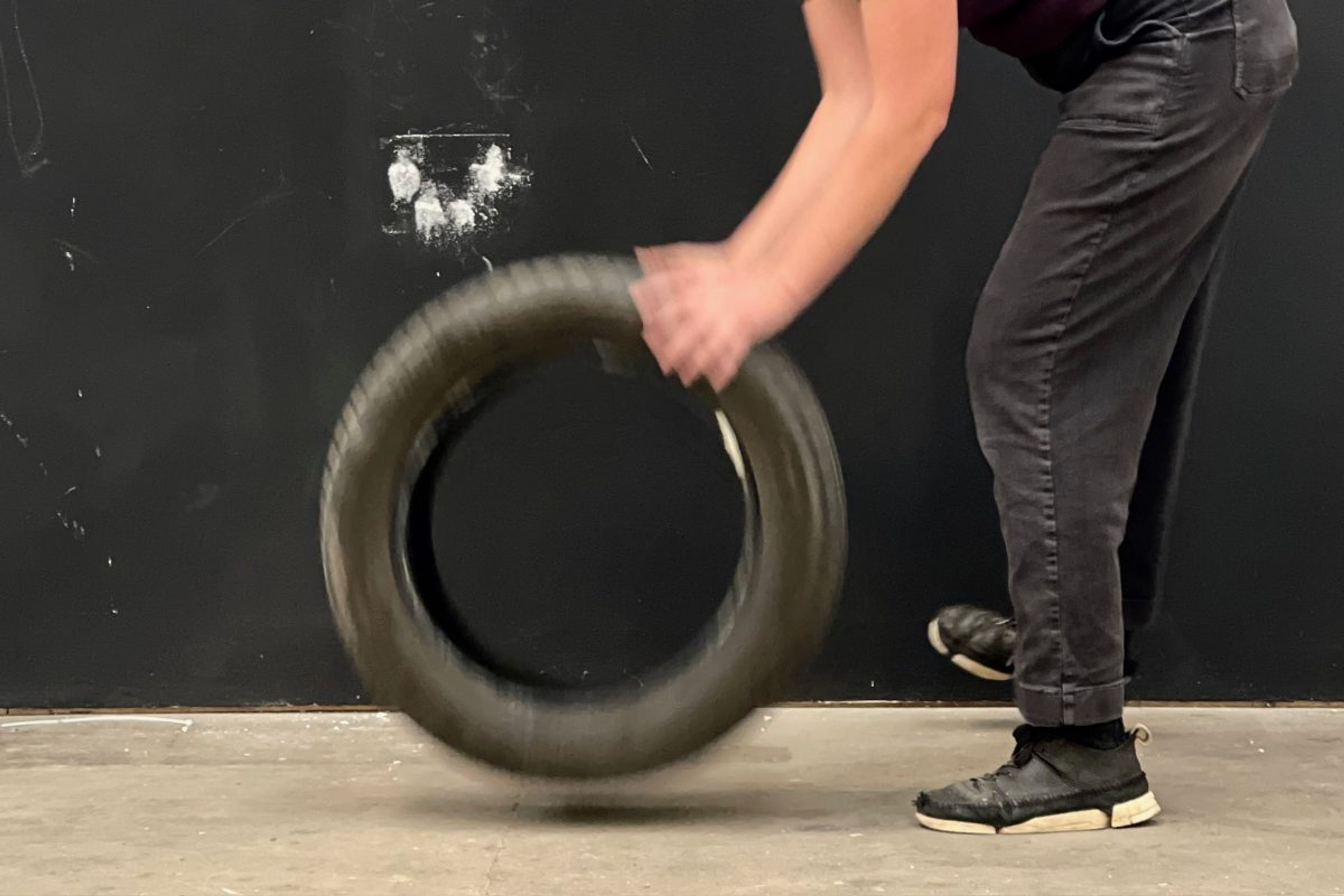 A person only visible from the torso downwards, wearing black jeans and black trainers, holding a black car tyre