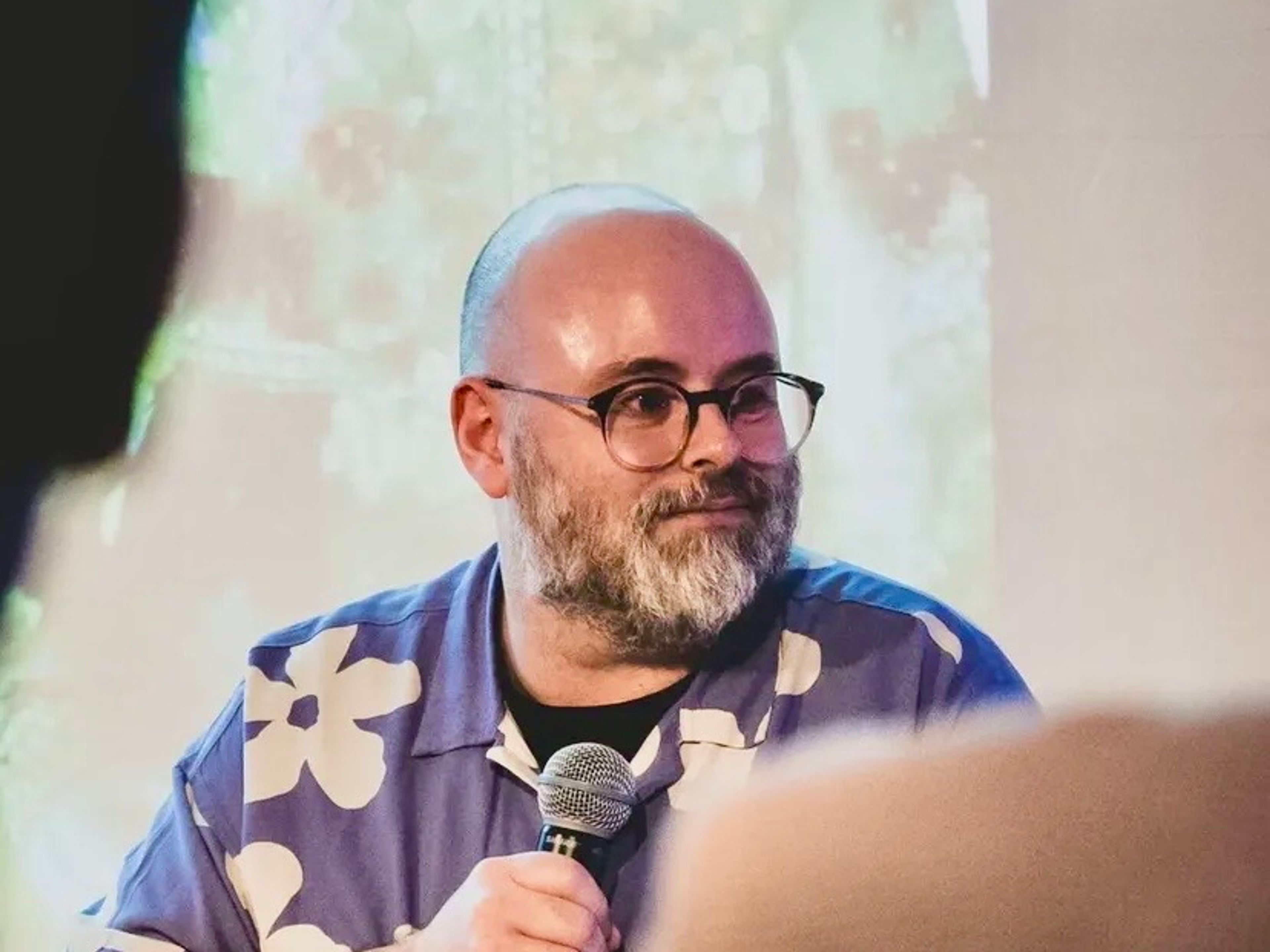 Kevin holding a microphone wearing glasses and a purple and white floral shirt.