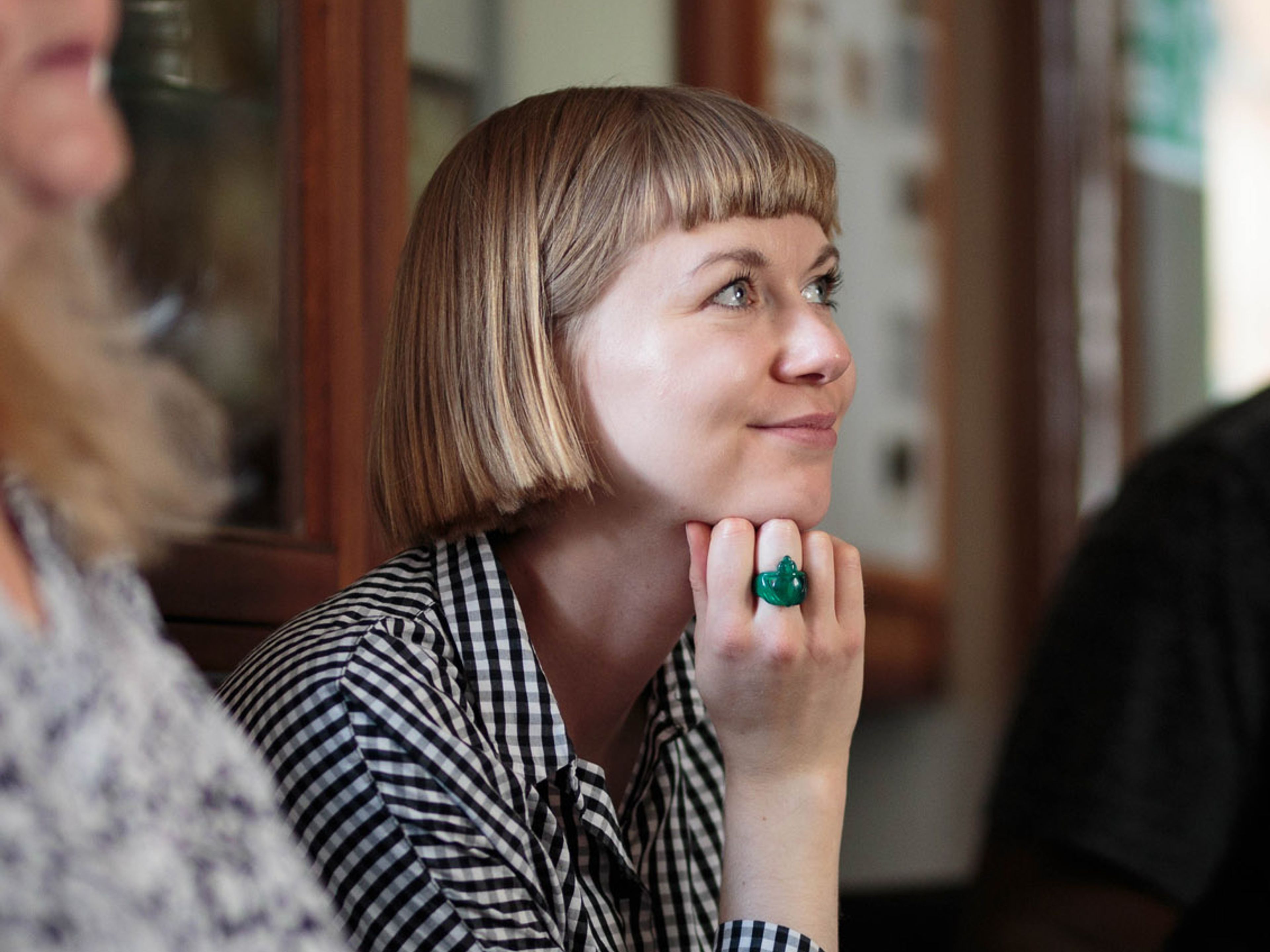 Colette is seated and stares to the right. She is wearing a gingham shirt and has a blonde bob.