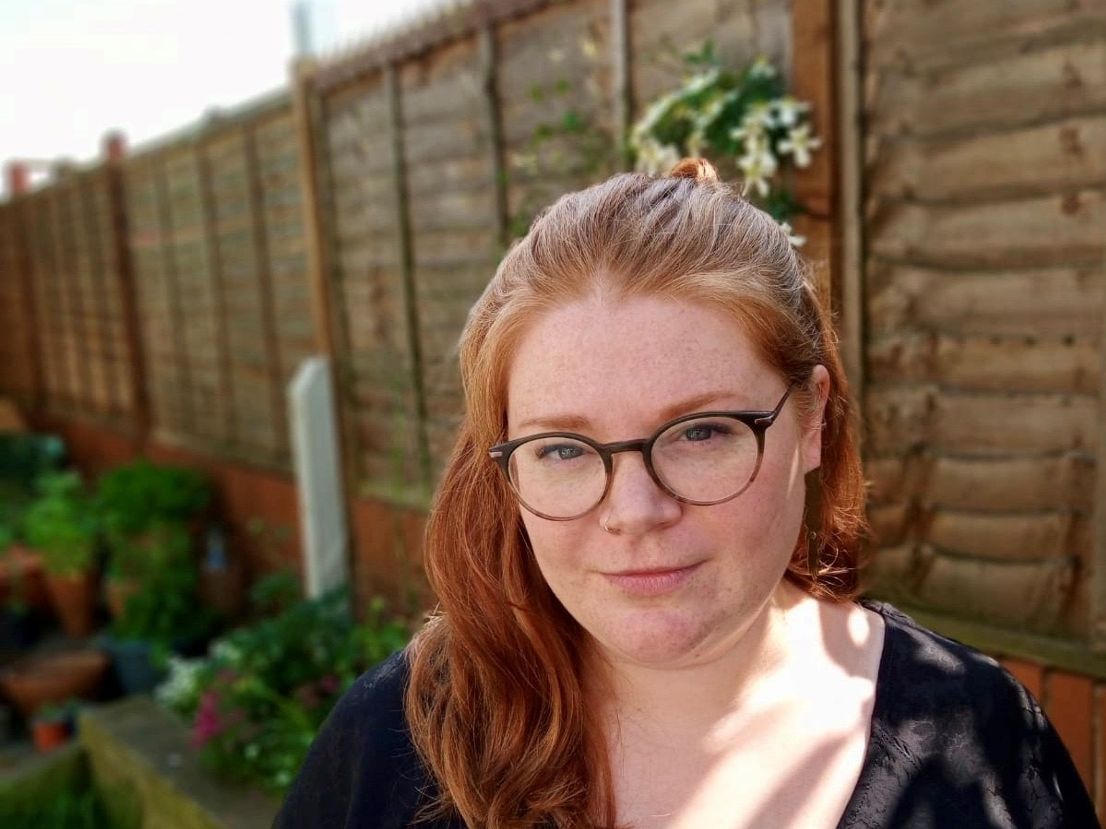 Lucy in a garden, they are wearing glasses and their ginger hair is just past shoulder length.