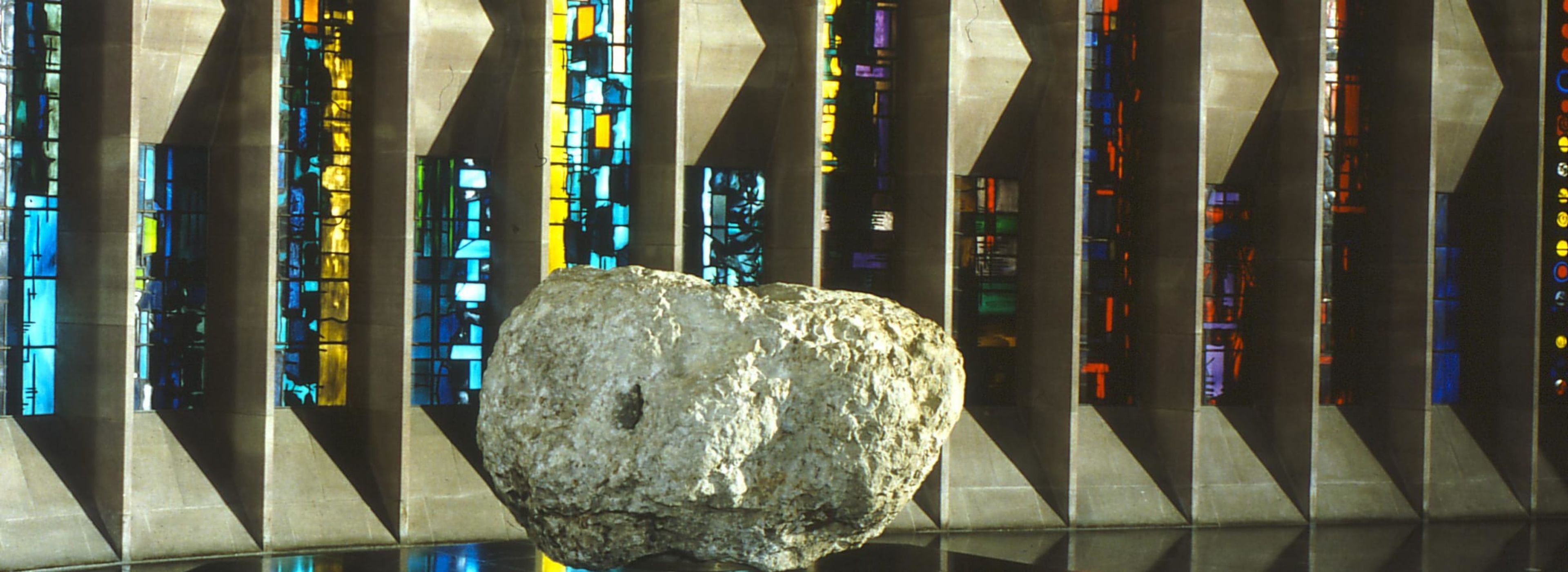 Large raw stone font, set indoors before tall stained‑glass windows, colourful geometric panels casting light onto the boulder and Cathedral floor.