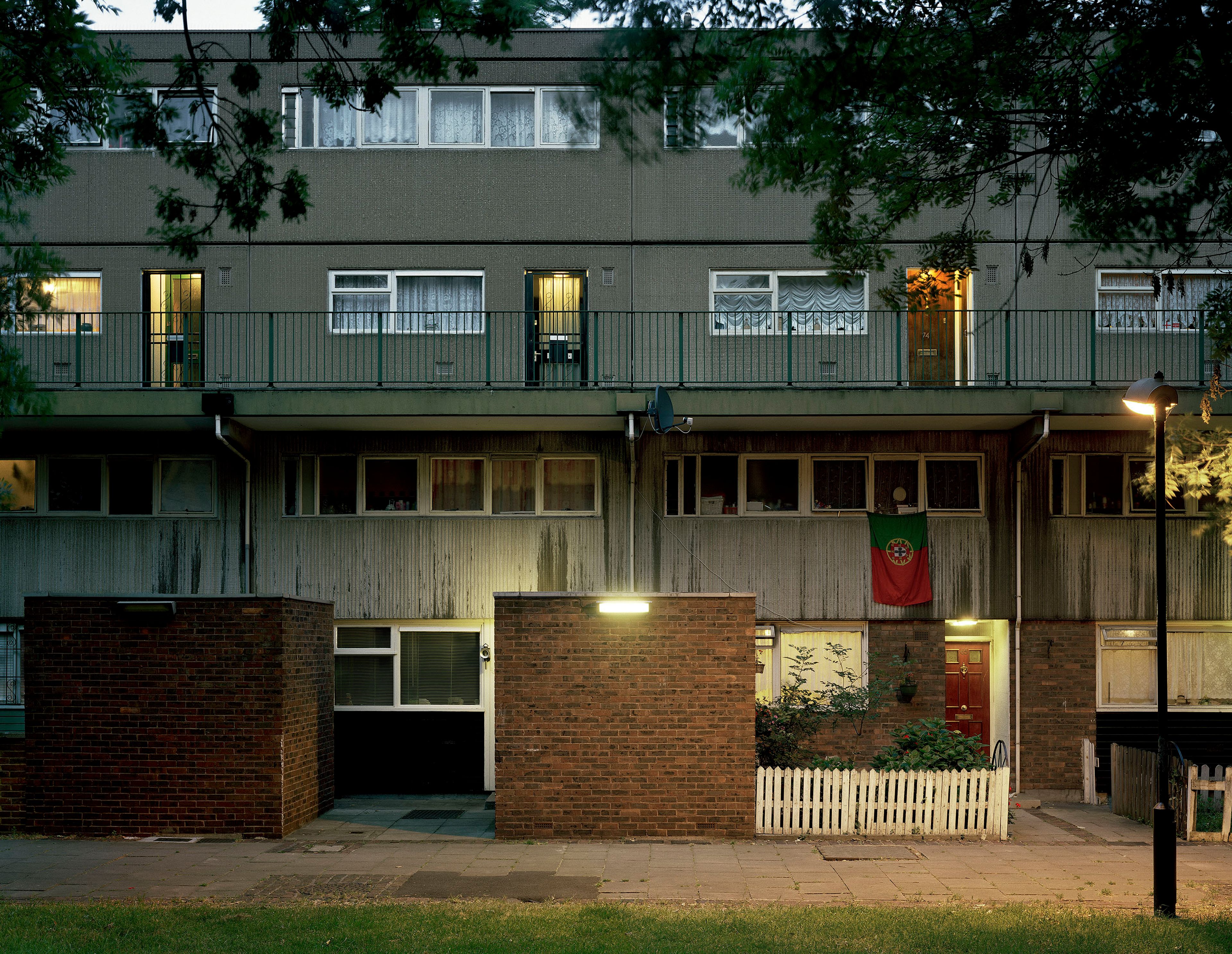 A two‑storey apartment building at dusk. Brick façades, small fenced 'garden' areas. Portuguese flag from one of the downstairs windows.