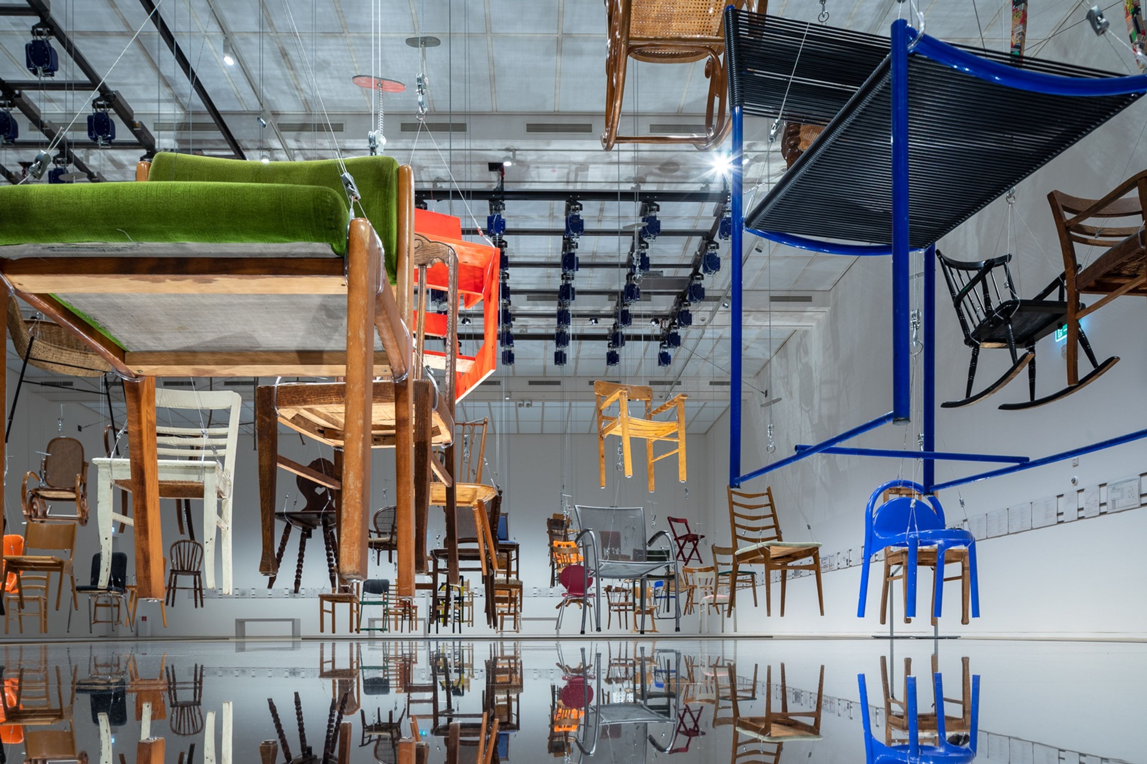 Large gallery installation featuring dozens of suspended chairs hanging at different heights above a reflective floor. 