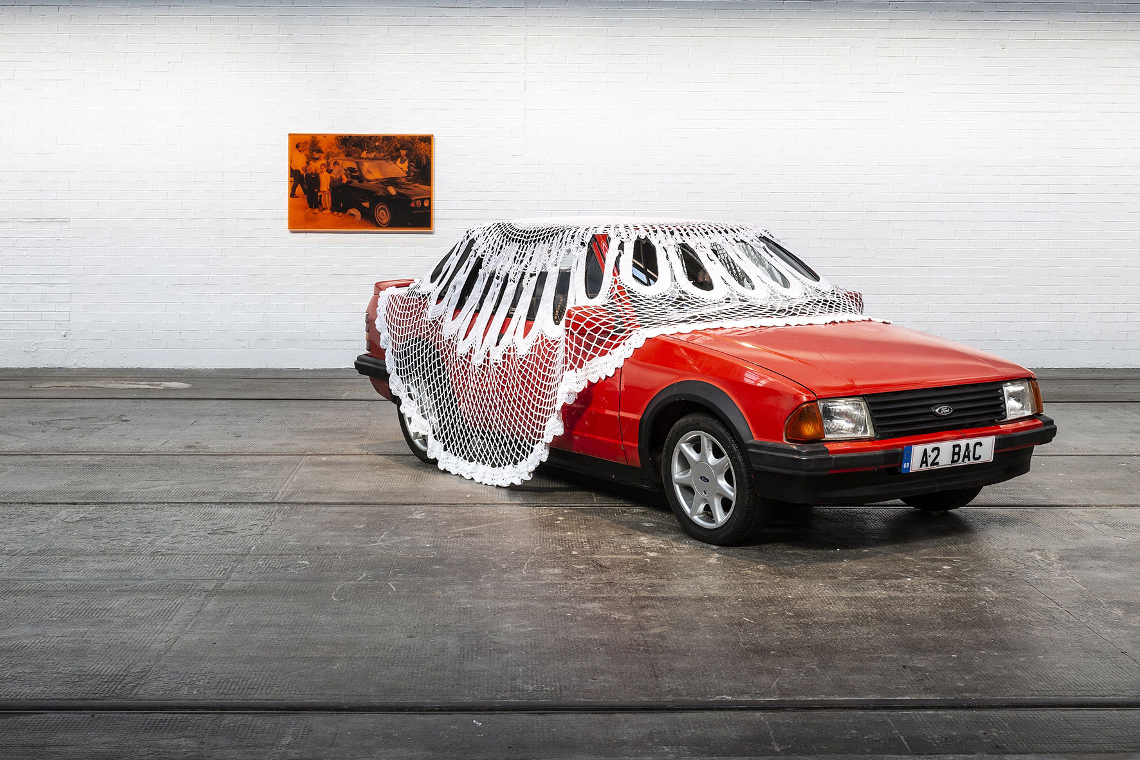 A red car is covered in a large outsized white doily in a white walled room on a grey concrete floor.