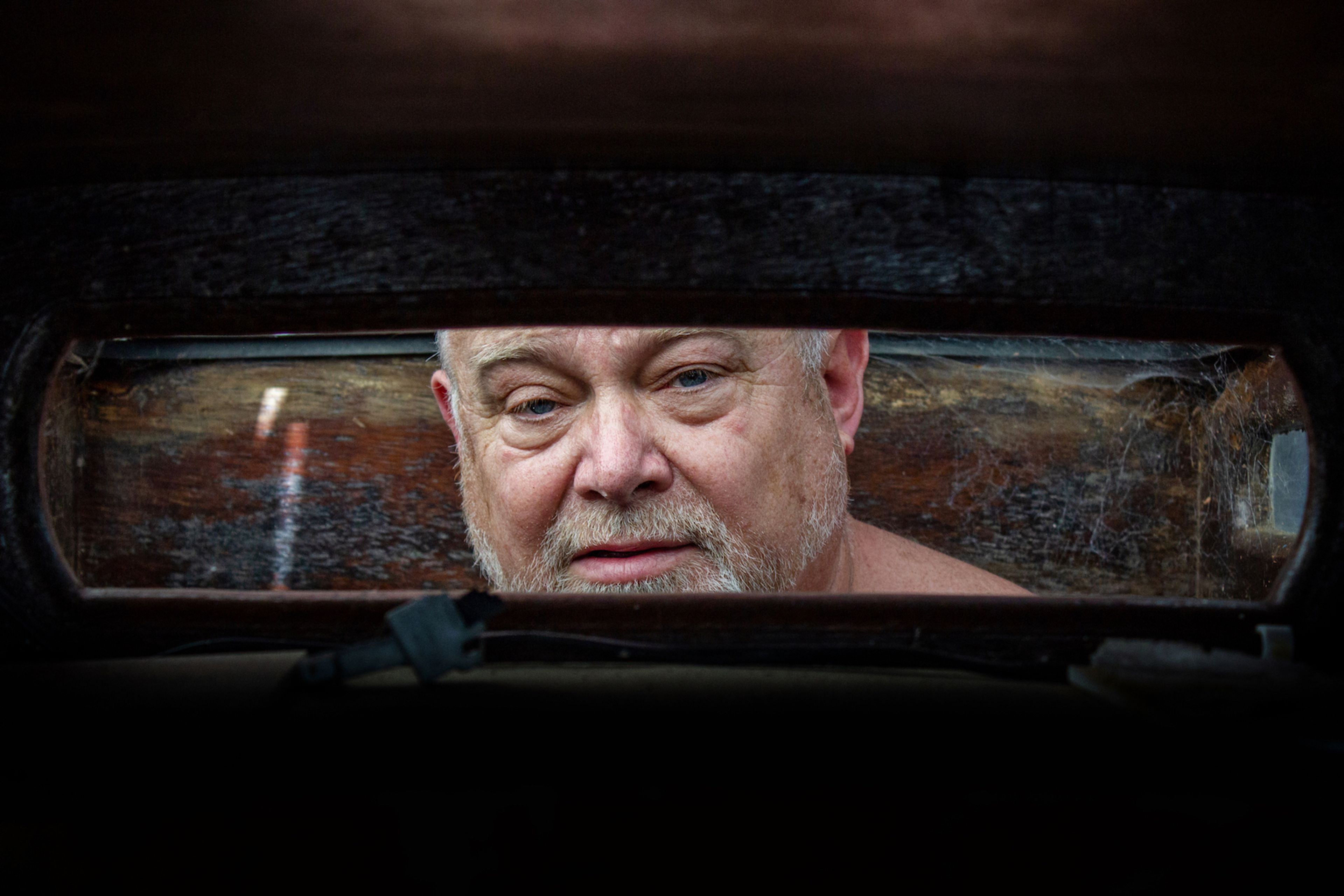 A photograph showing a man peering through a boat window. His face is framed by the window, and is the main focus of the image. 