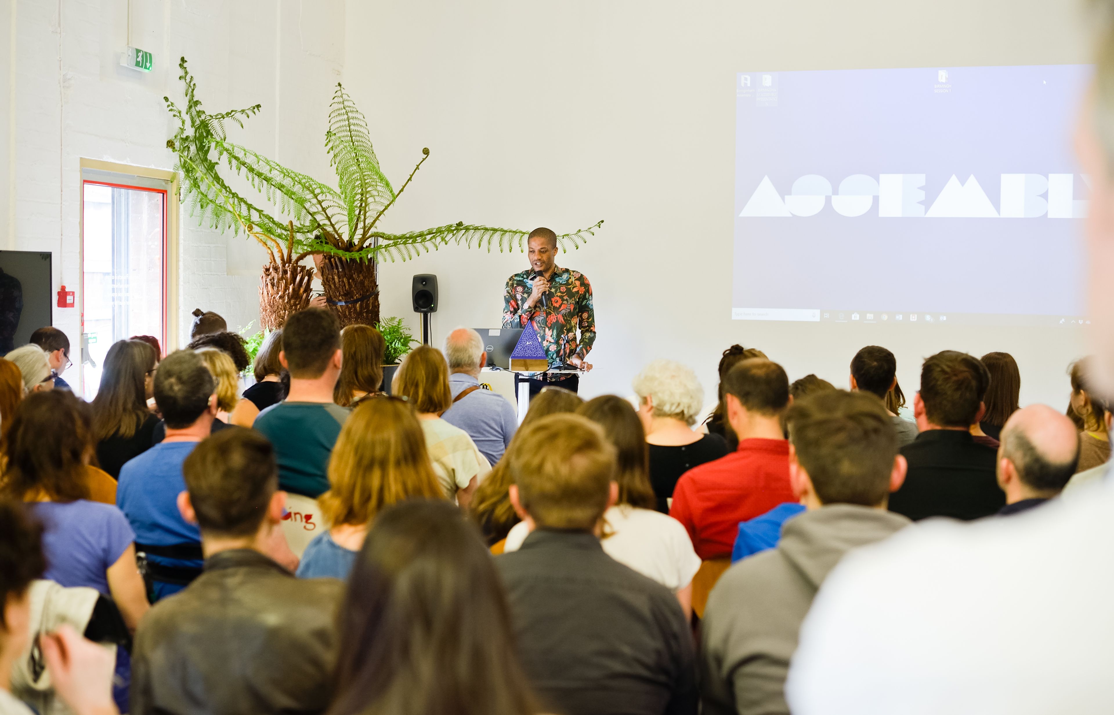 A crown of  seated individuals are addressed by a speaker in front of a screen that reads Assembly.