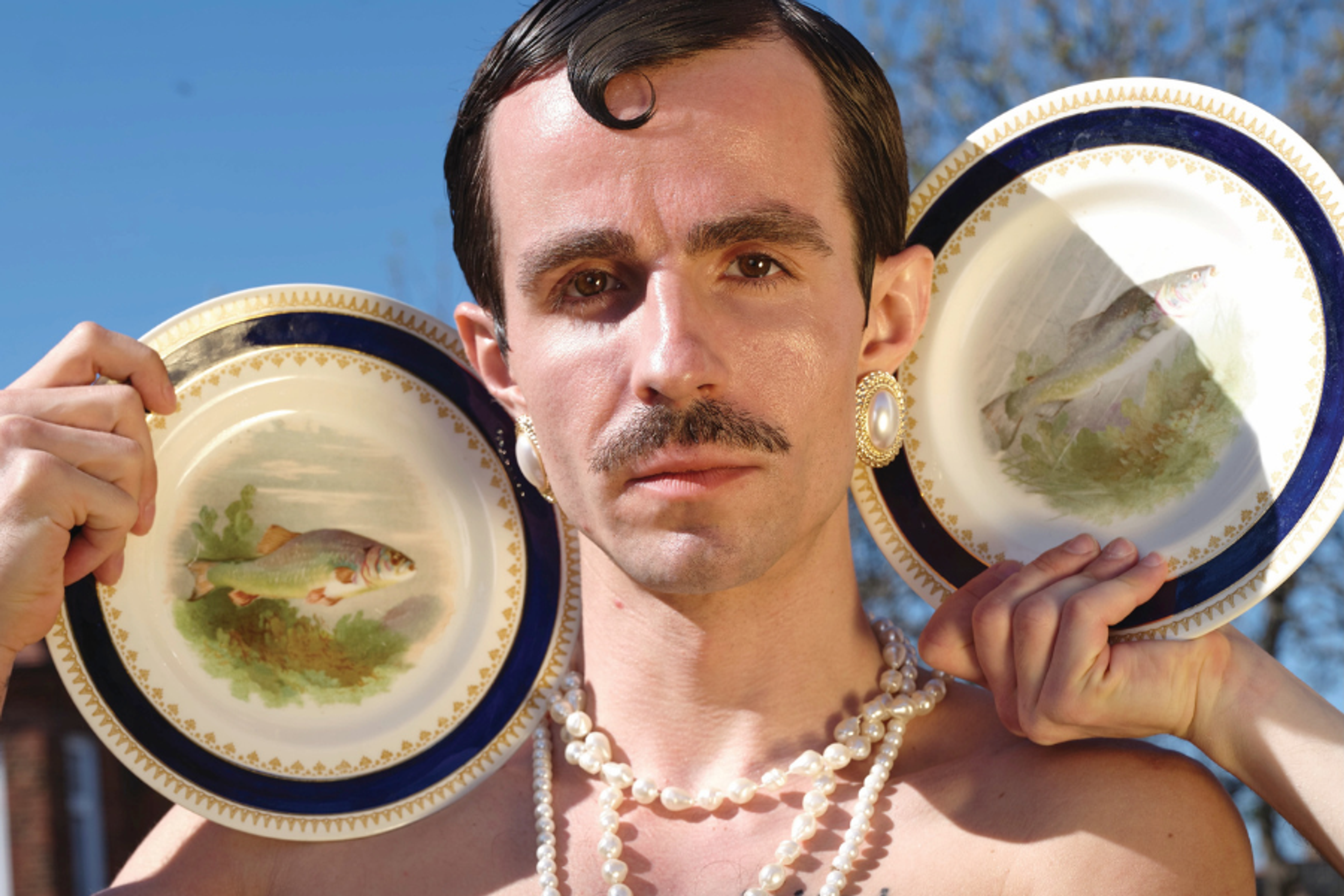 The photo presents a person posed of Queer & Eastern European identity, wearing regal-style pearl necklaces and earrings, whilst holding up a plate.