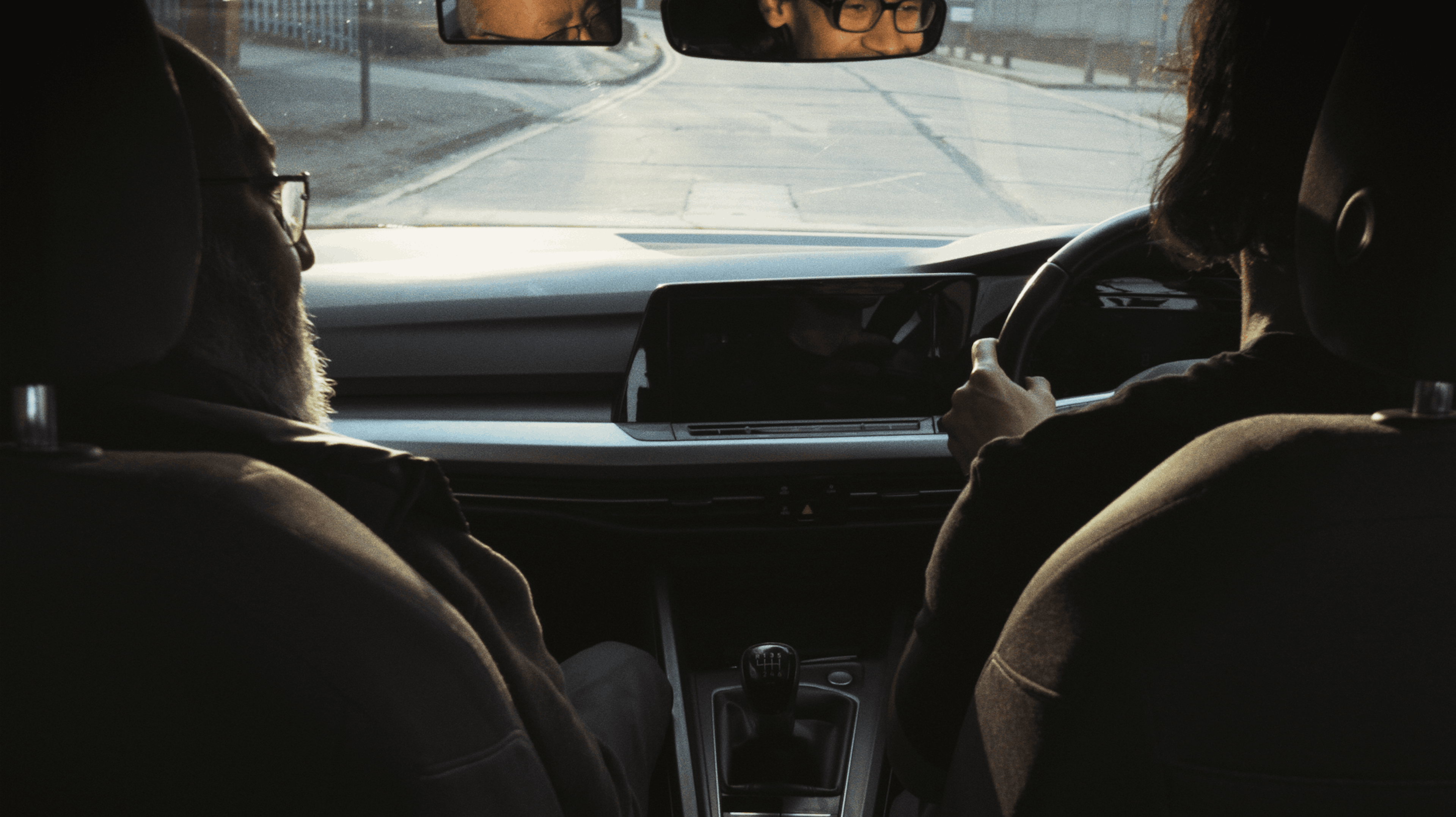 Two figures are sat together in a car. The passenger is looking to the driver. The driver is smiling in the rear view mirror.
