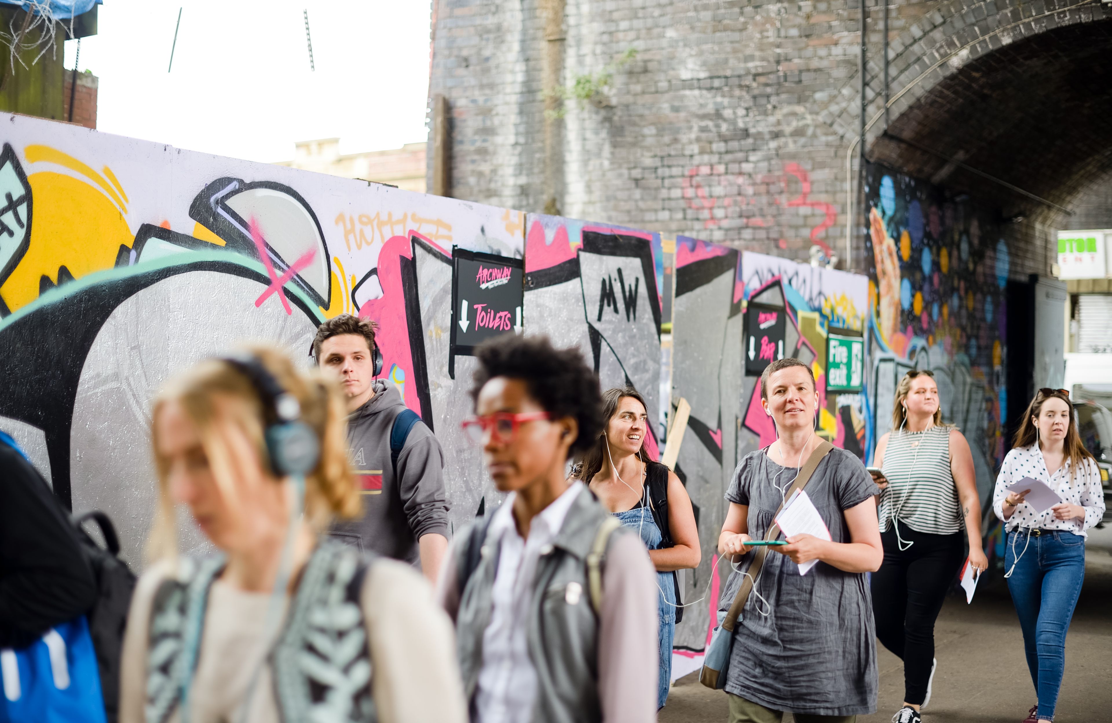 A group of people walking under a bridge and in front of a graffiti art wall.