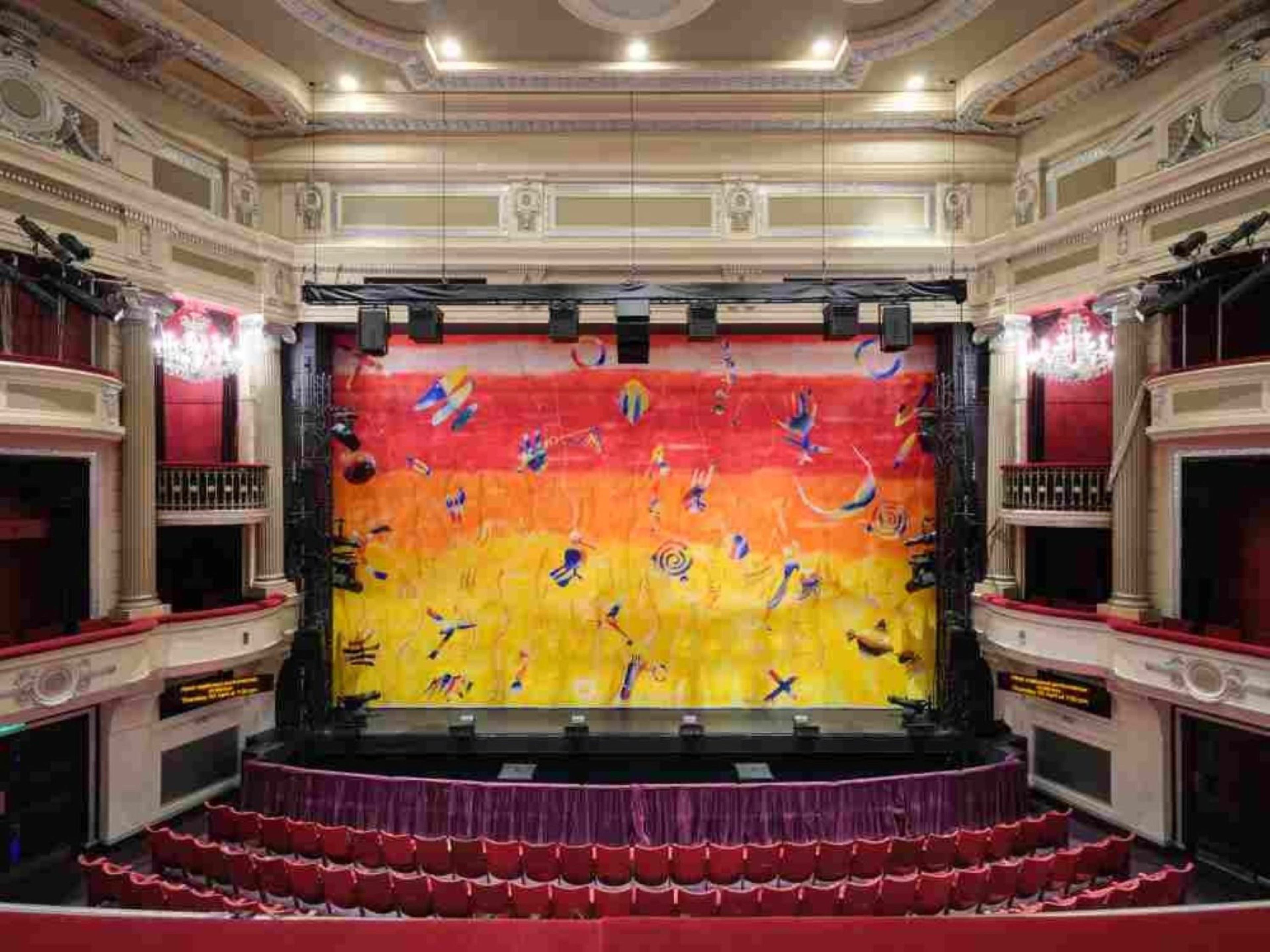 Theatre auditorium with red seats, ornate balconies anf chandeliers, facing a stage framed by a vibrant red-to-yellow abstract curtain.