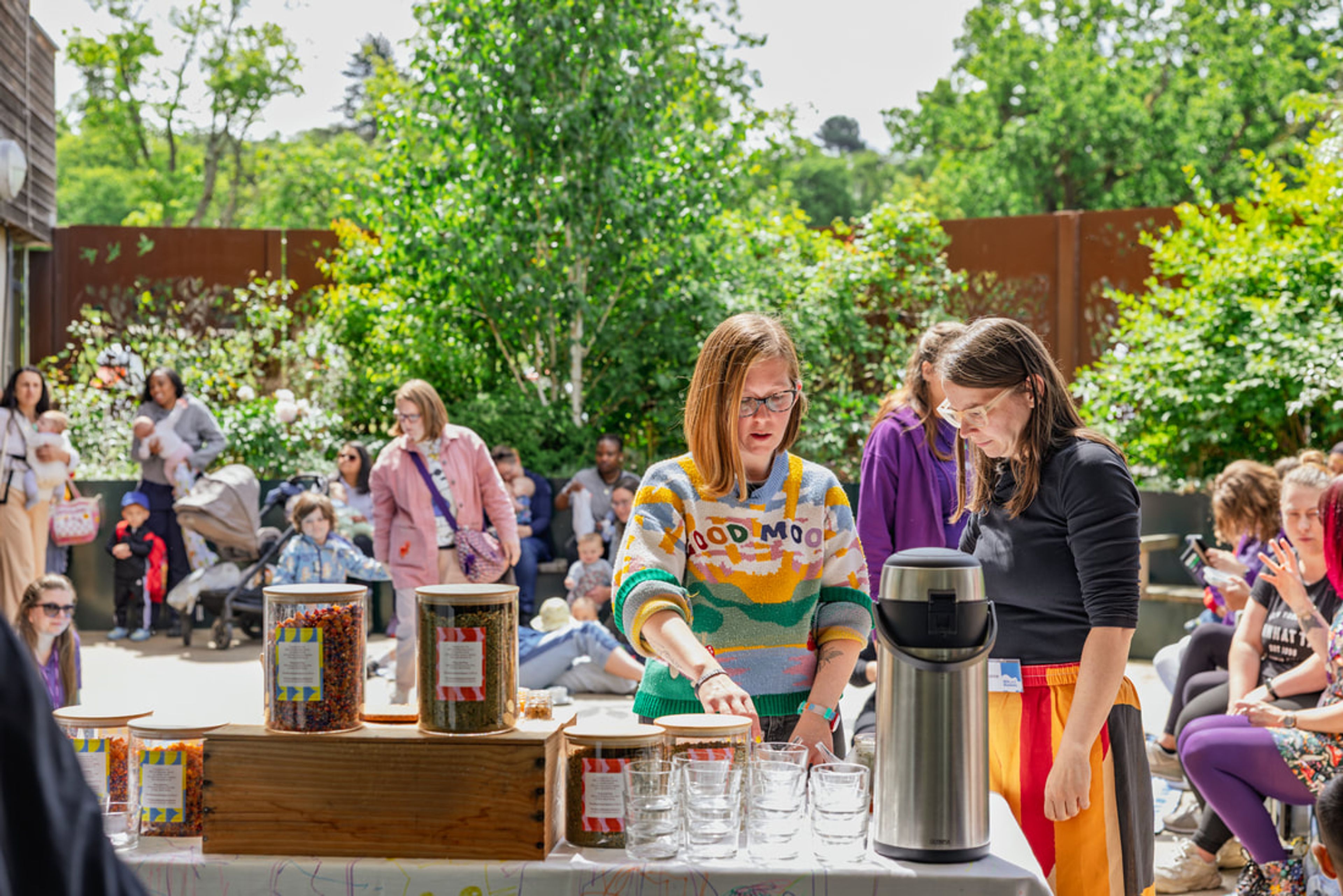 Amelia Hawk serving drinks at an outdoor event, standing behind a table with glasses and jars, while families gather in a sunny garden setting behind. 
