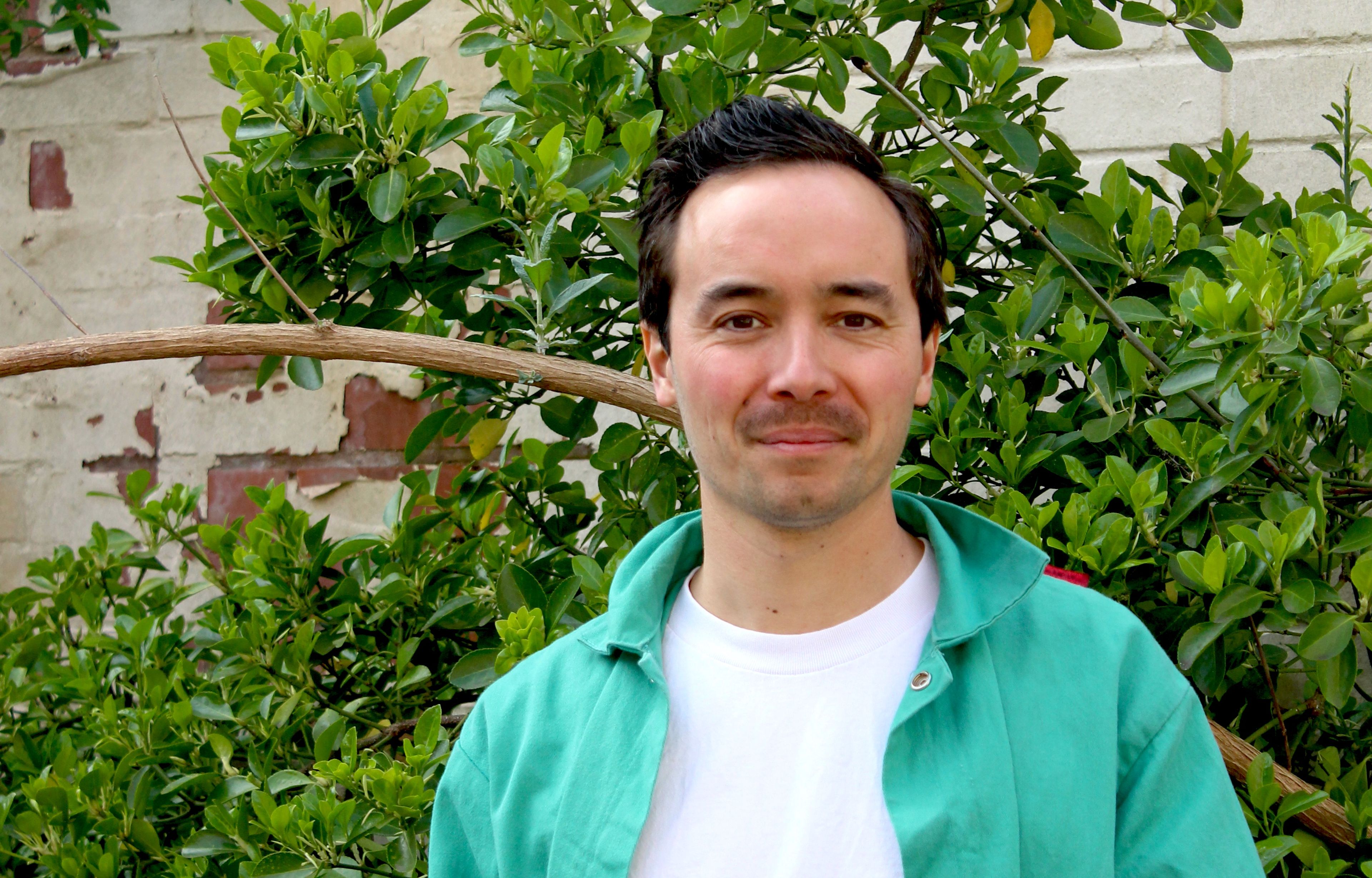 Portrait of John against foliage. John is wearing a white t-shirt and green shirt.