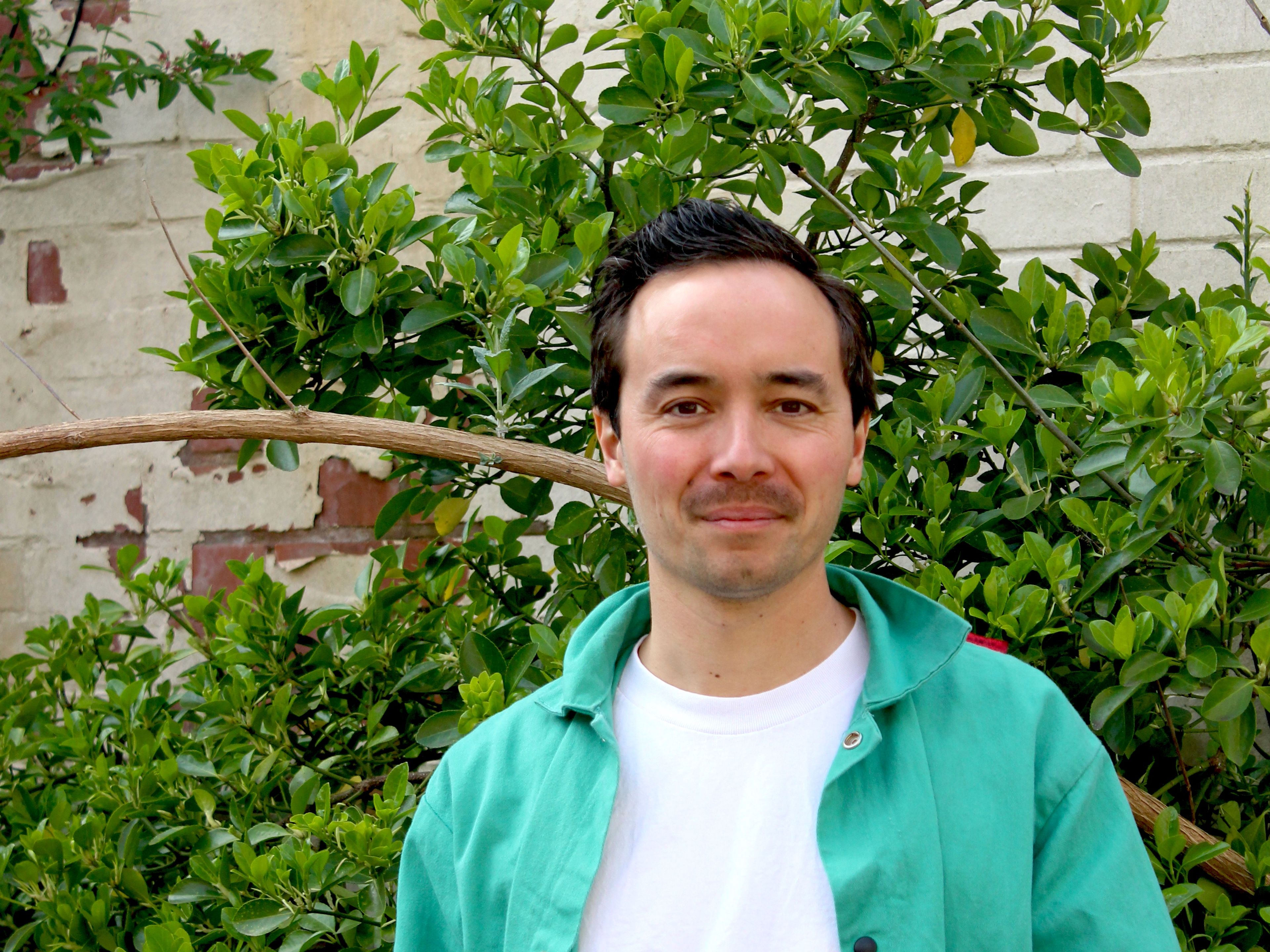 John is wearing a white t-shirt and green shirt. He is stood in front of lush foliage.