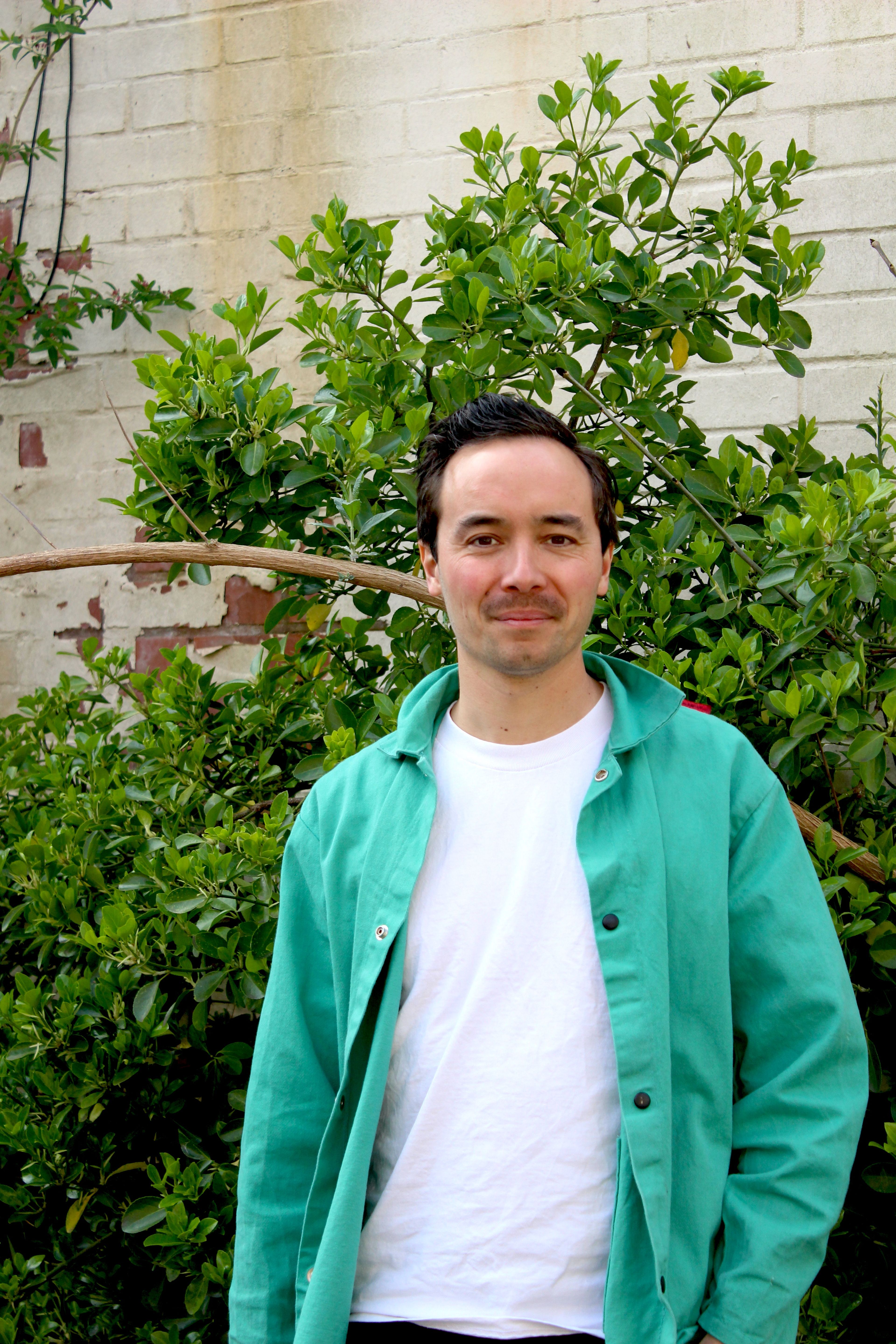 Portrait of John against foliage. John is wearing a white t-shirt and green shirt.